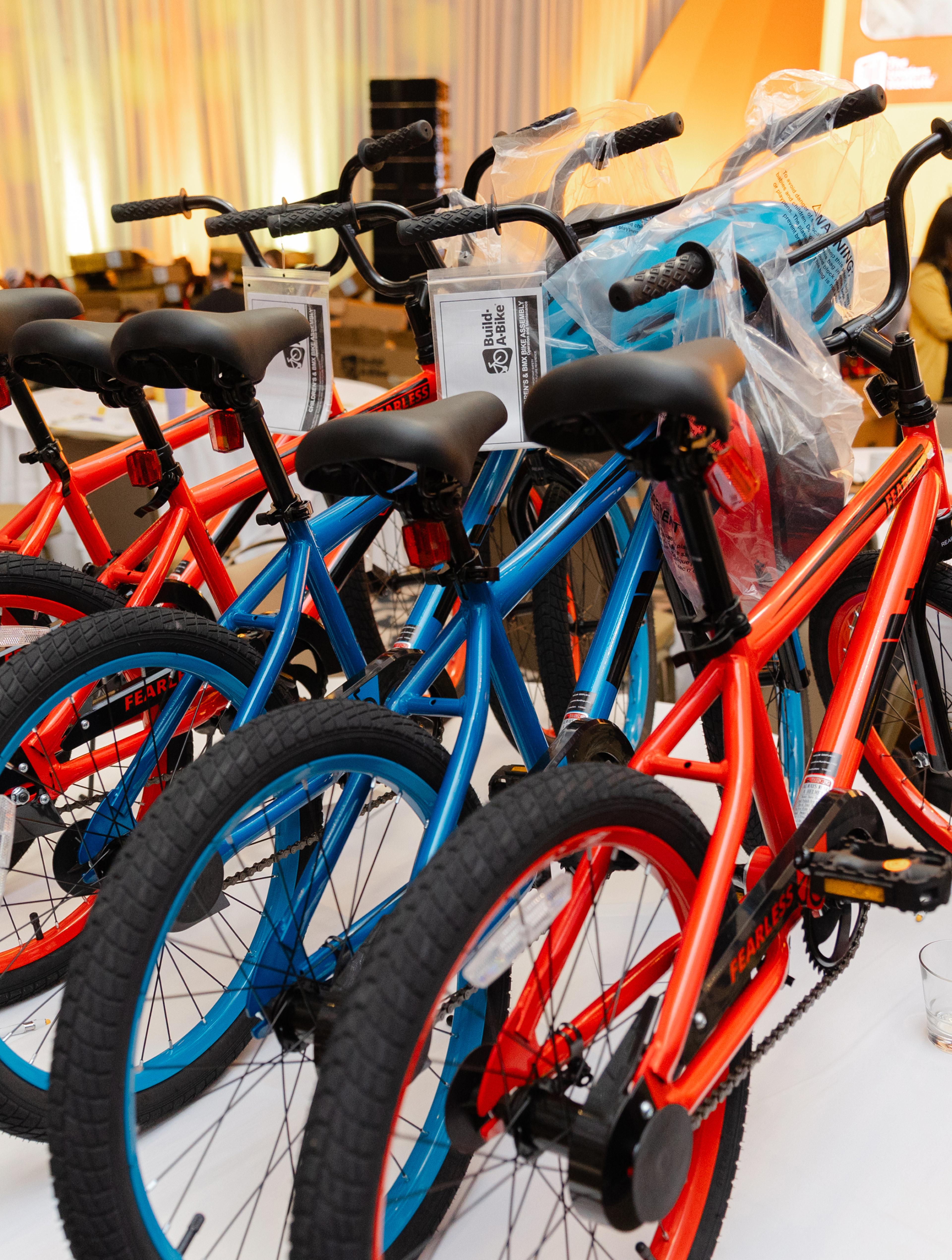 Rows of red and blue bicycles lined up indoors, ready for donation or assembly, with “Build a Bike” tags attached to the handlebars.