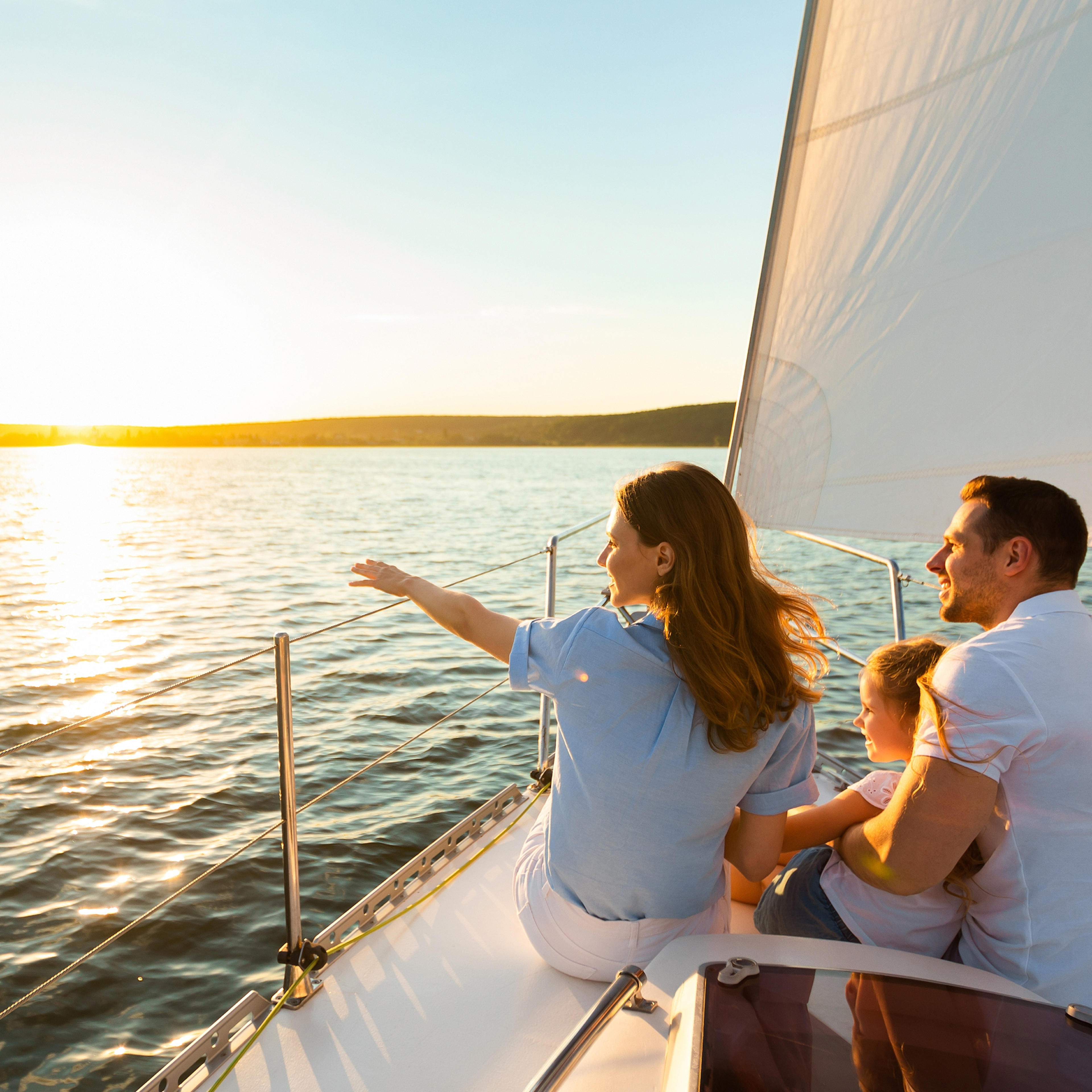 Family of three sailing on yacht sitting on sailboat deck looking at sunset at seaside.