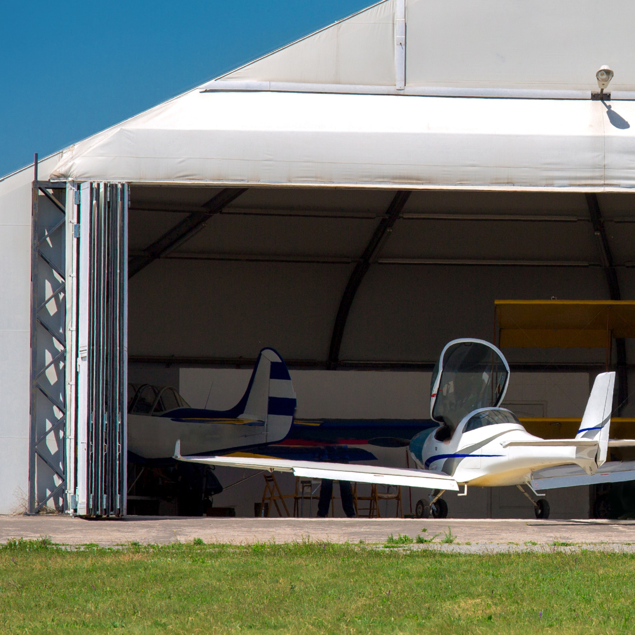 Two small aircraft are parked inside an open hangar on a sunny day, with green grass visible in the foreground.
