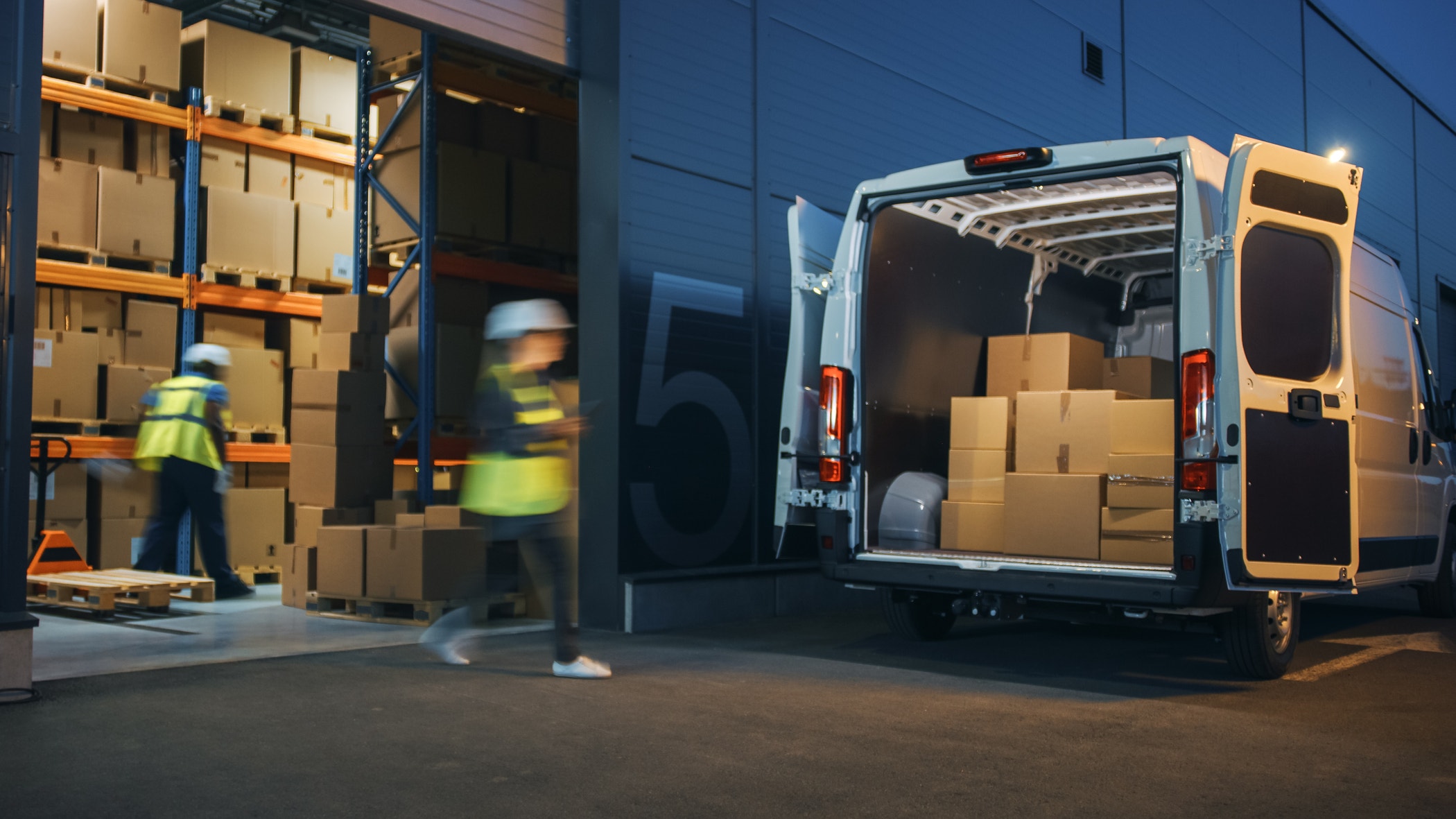 Outside a logistics distribution warehouse, a diverse team of workers loads a delivery truck with cardboard boxes for online orders and e-commerce goods.