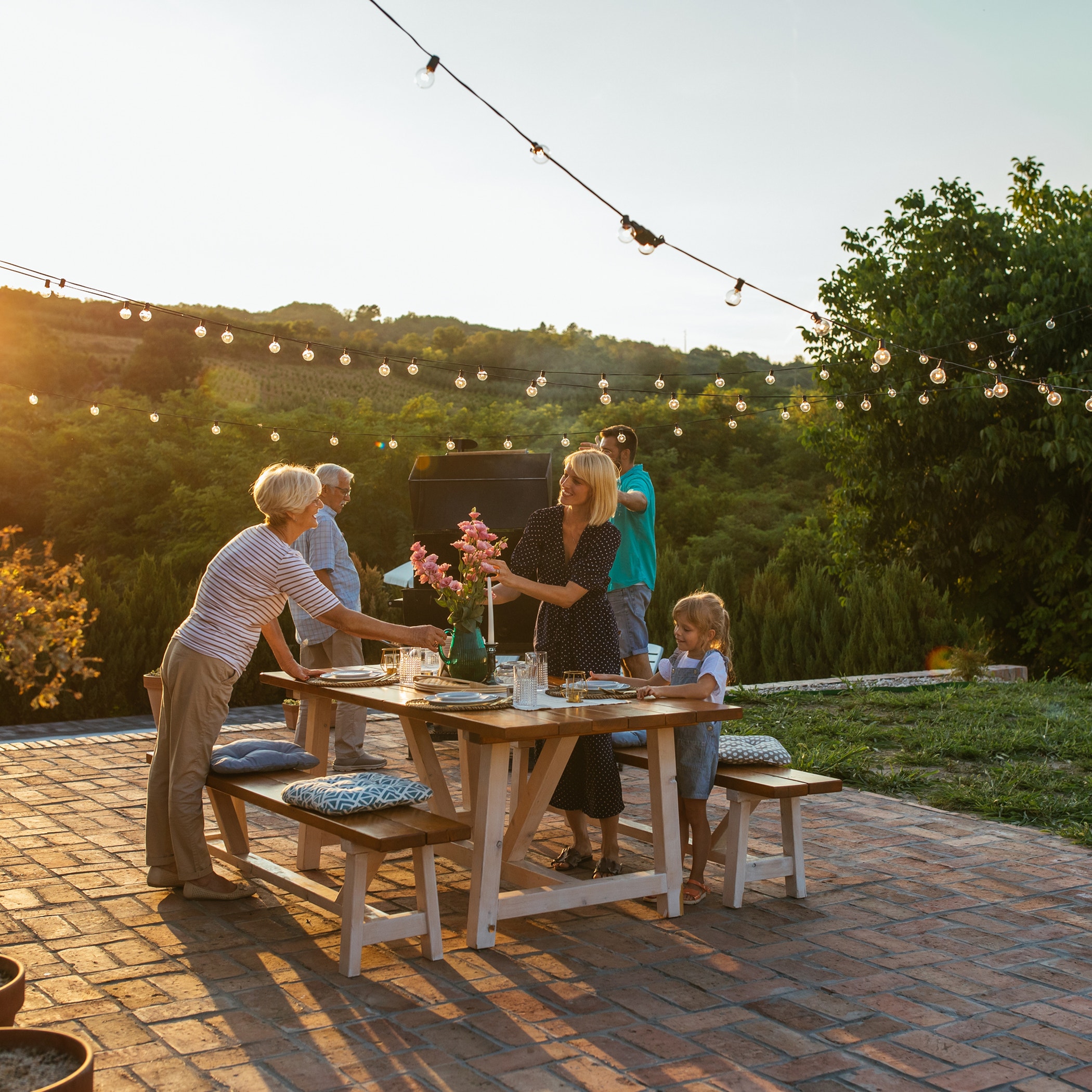 A family gathers around an outdoor wooden table set for a meal, with string lights overhead and greenery in the background at sunset.