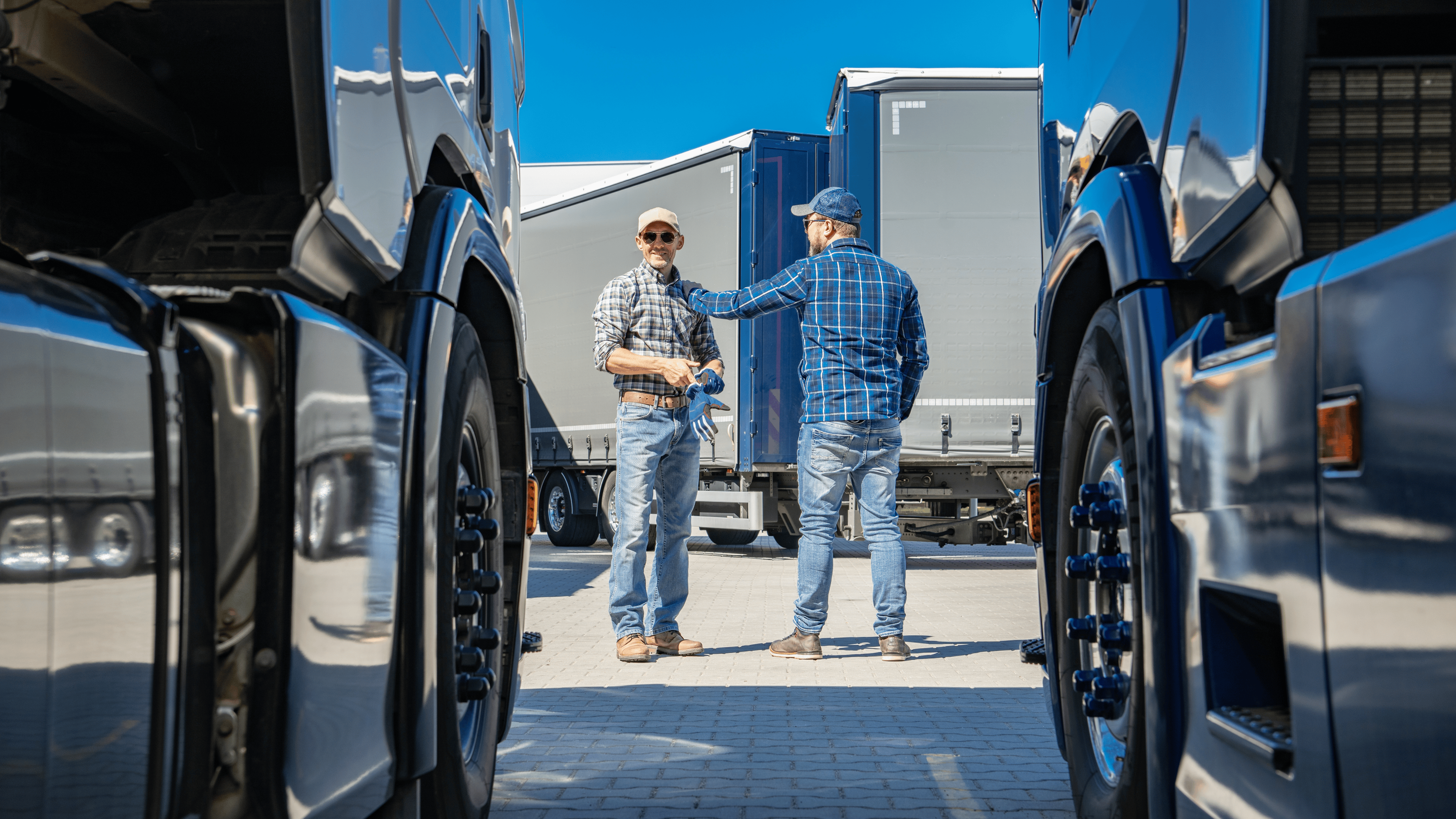Two truck drivers talking about logistics between parked trucks in a busy transport yard under a bright blue sky.