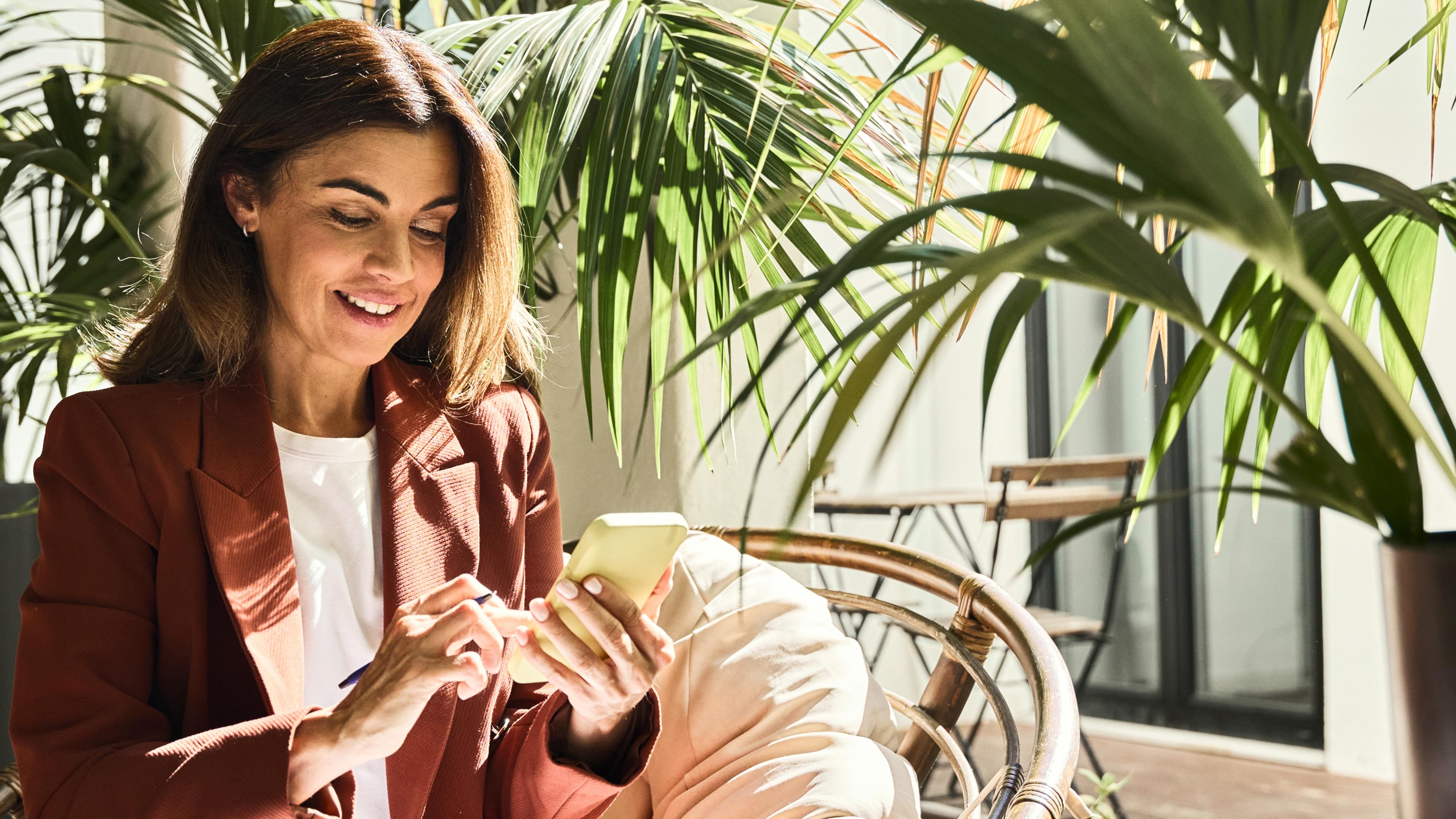 Happy smiling elegant mature middle aged business woman wearing suit holding cellphone using mobile cell phone looking at smartphone sitting in comfortable chair in sunny office with green plants.