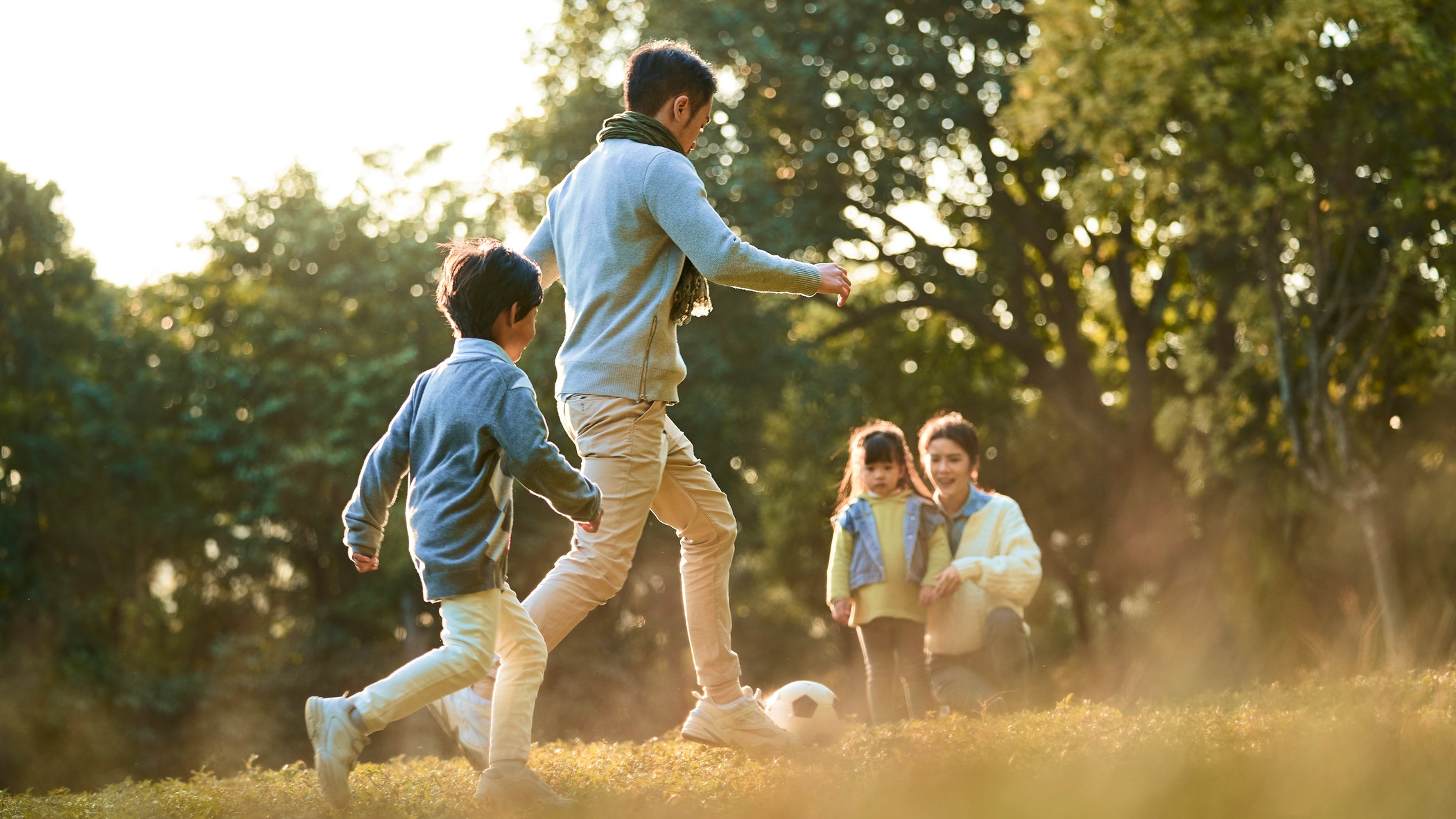 An adult and two children play soccer on grass while another adult and child watch nearby; trees and sunlight are in the background.