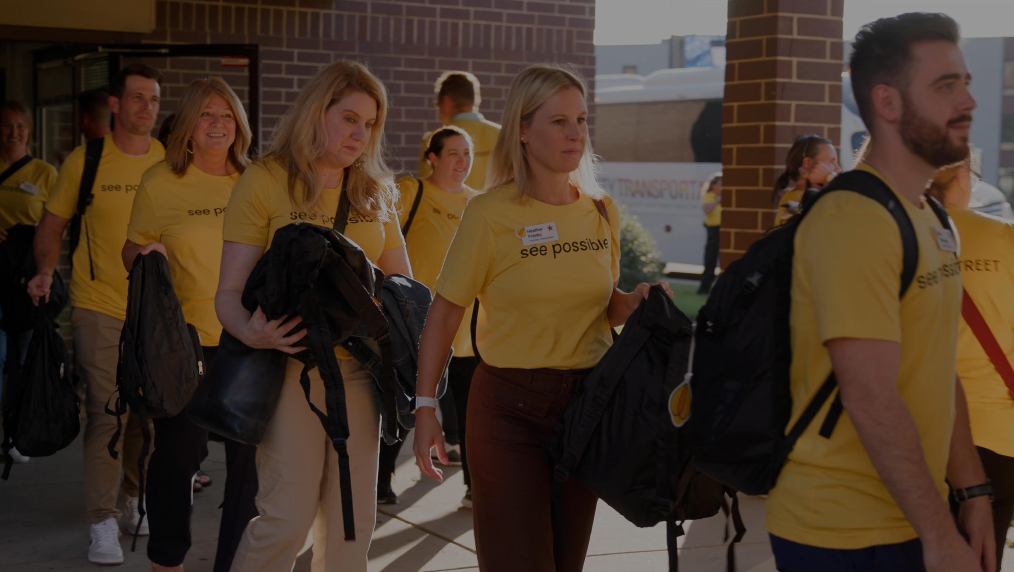 Woman walking with a group wearing a yellow see possible t-shirt carrying a backpack.