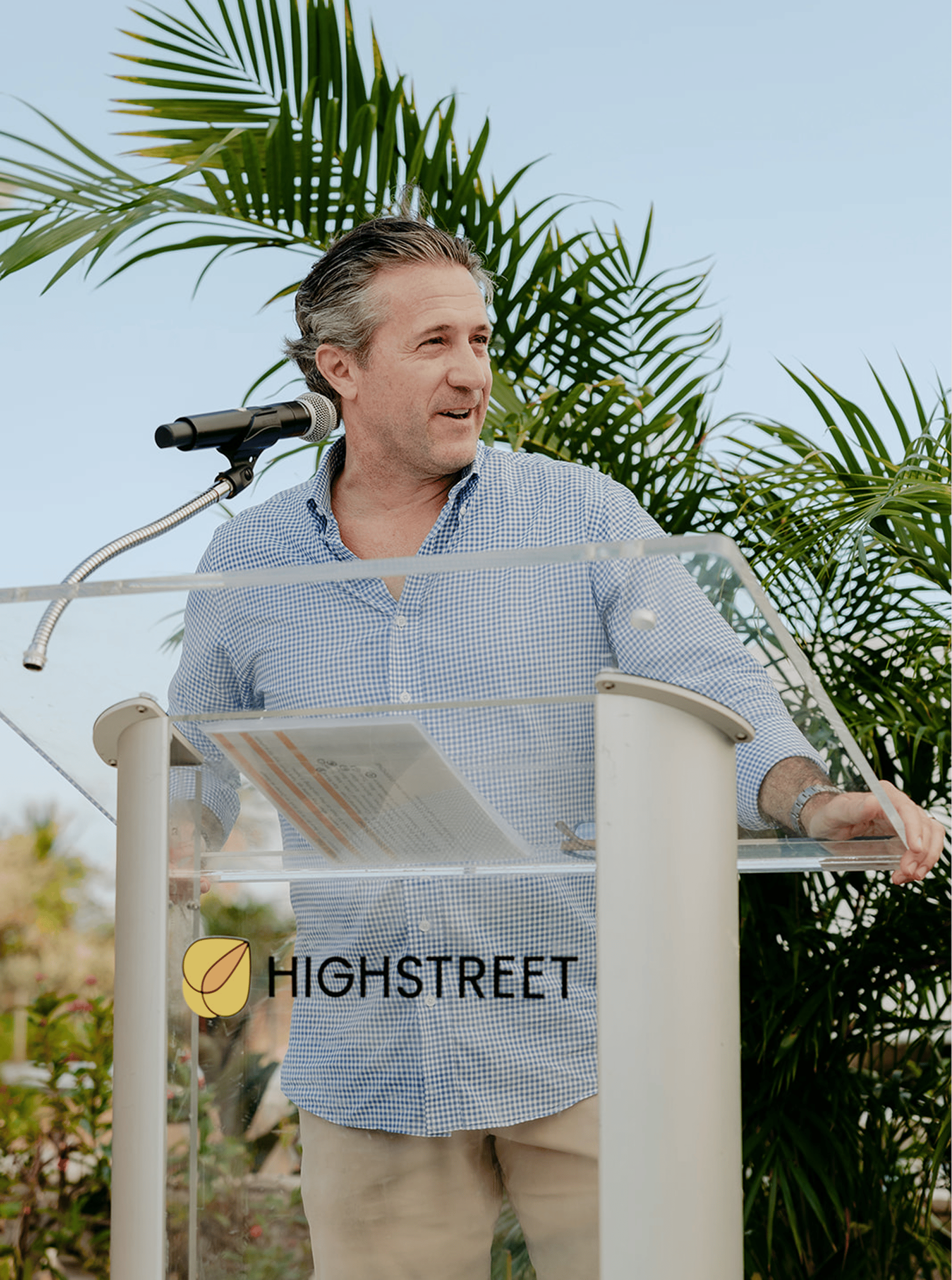 Man speaking outdoors at a podium with the Highstreet logo, surrounded by palm trees and bright daylight.