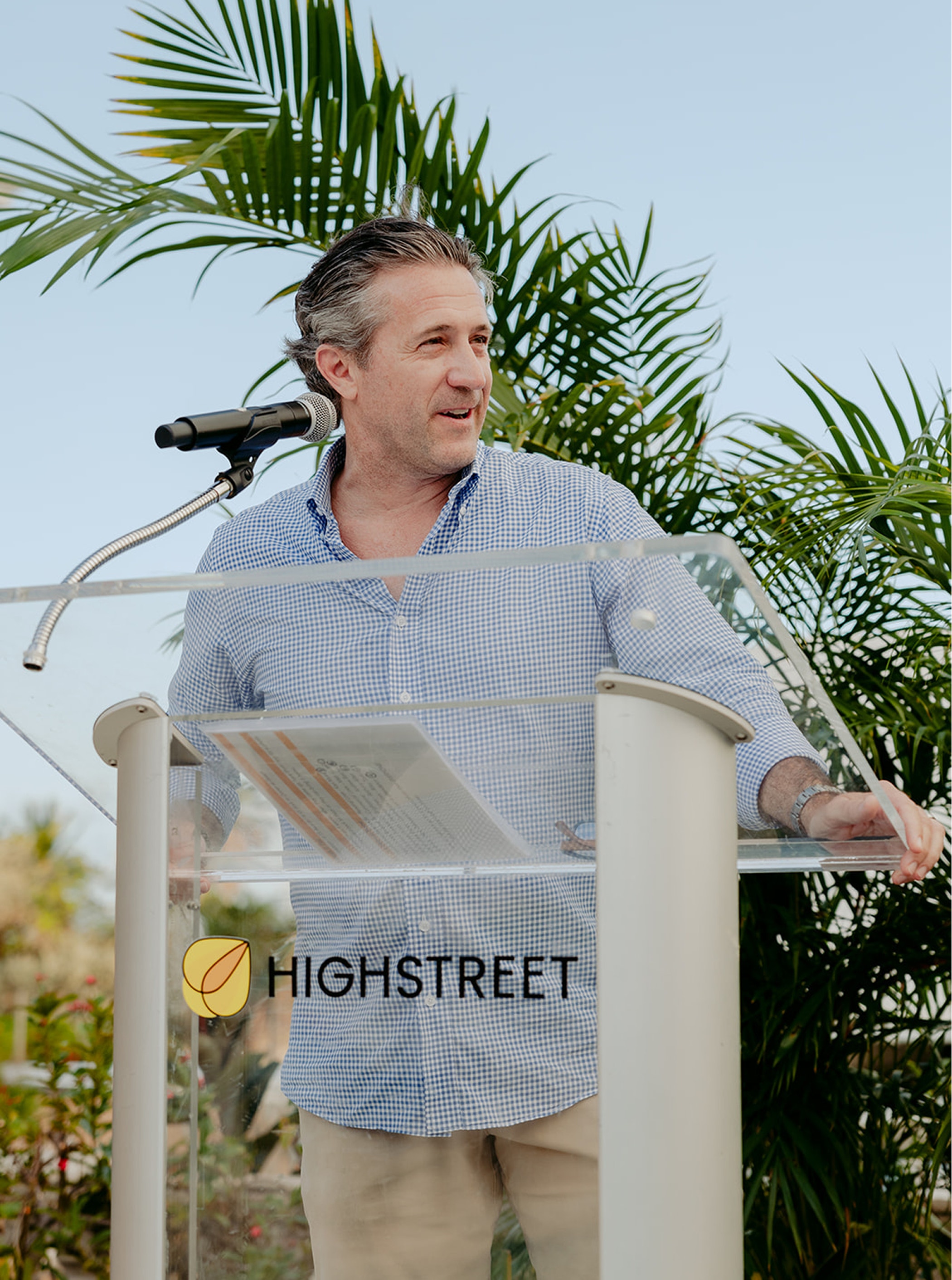Man speaking outdoors at a podium with the Highstreet logo, surrounded by palm trees and bright daylight.