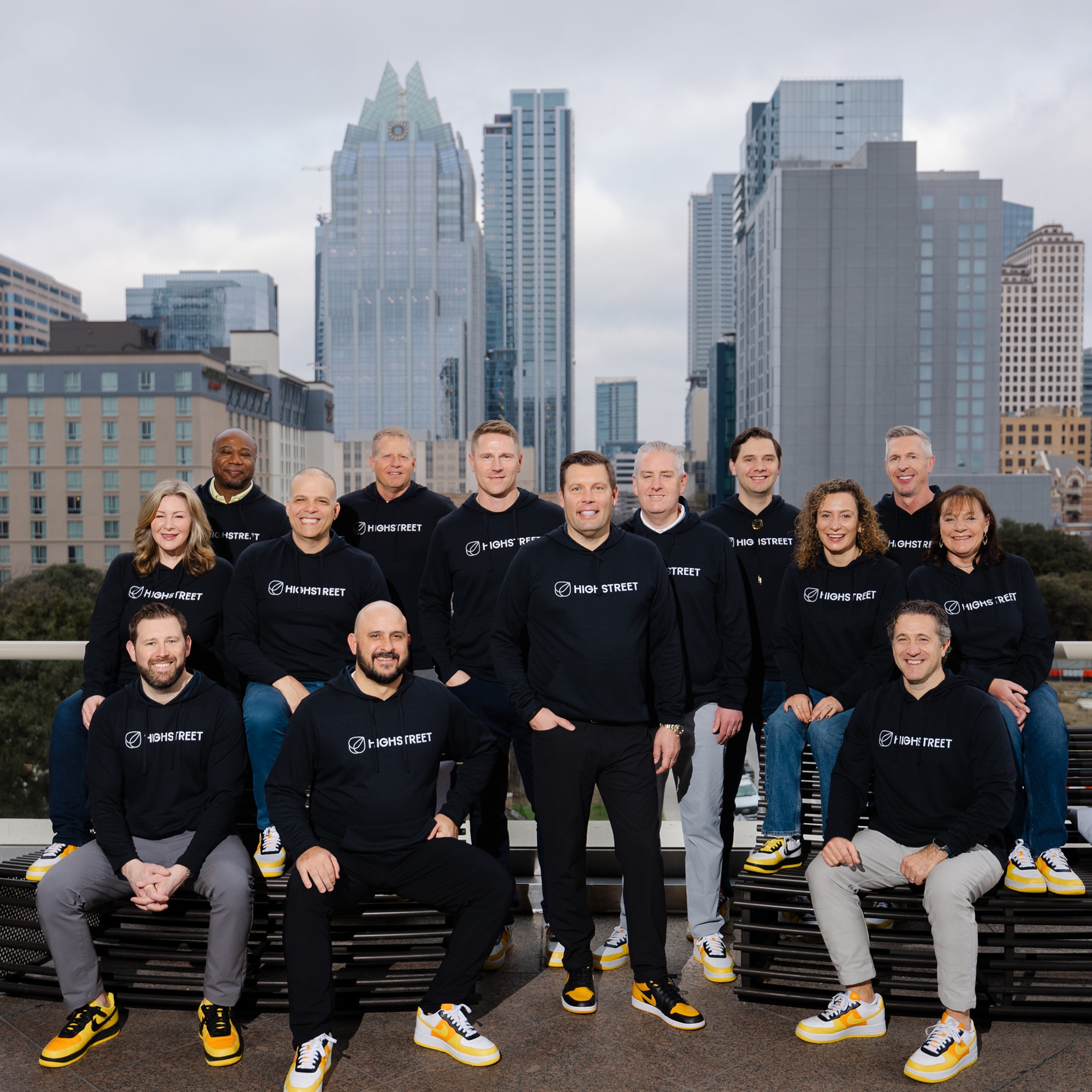 Group of people wearing black Highstreet sweatshirts posing together outdoors with city buildings in the background.