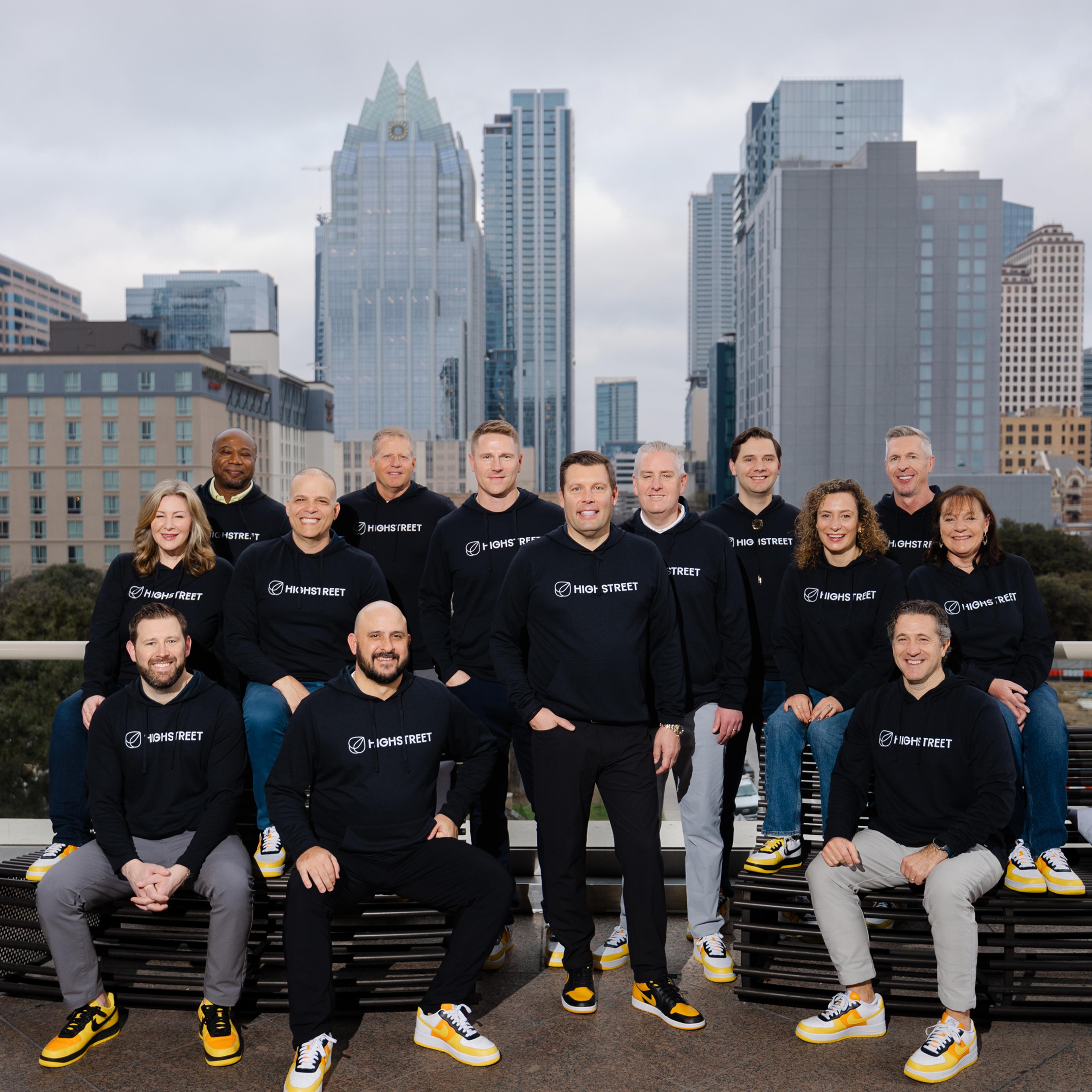 Group of people wearing black Highstreet sweatshirts posing together outdoors with city buildings in the background.