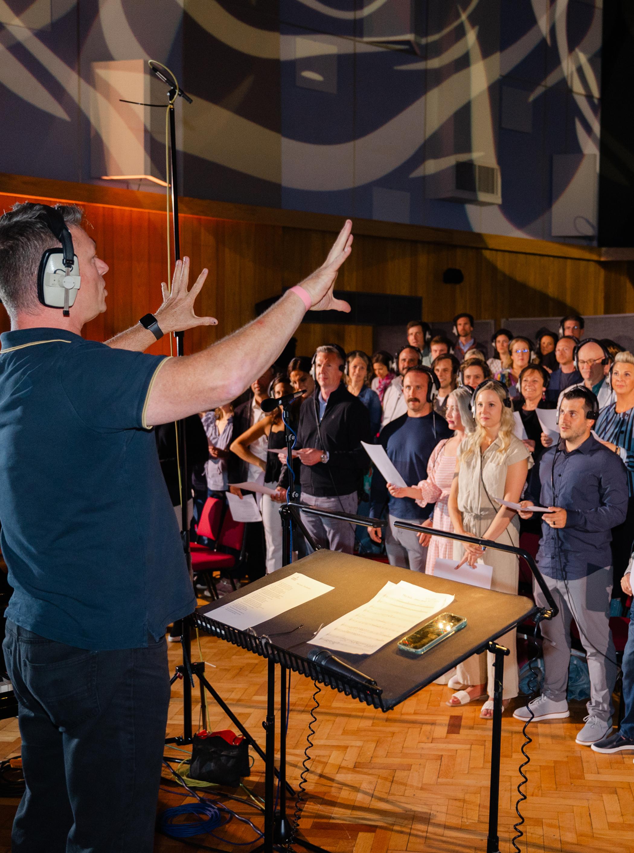 A conductor wearing headphones directs a choir in a recording studio. The singers hold sheet music and stand attentively.