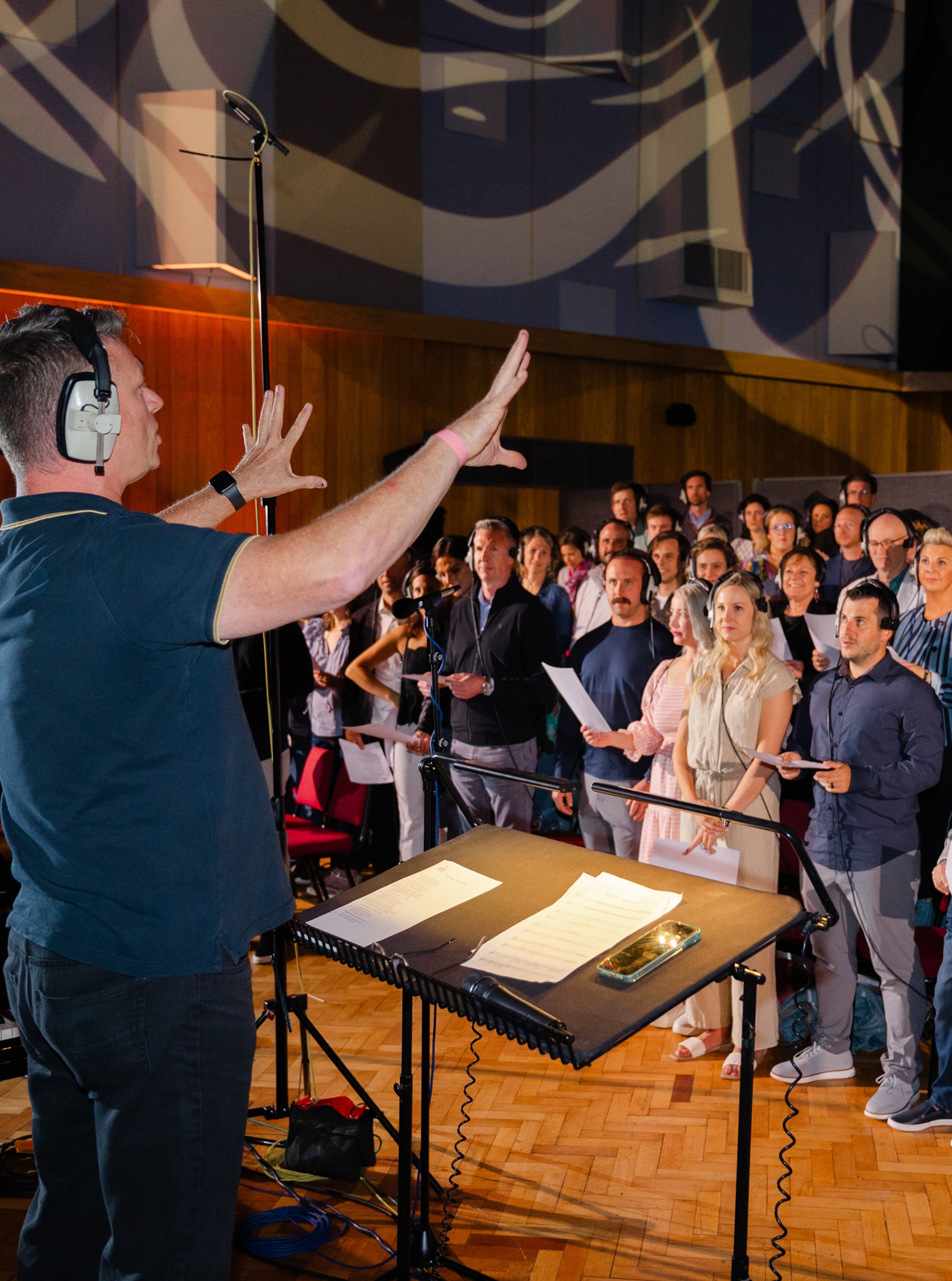 A conductor wearing headphones directs a choir in a recording studio. The singers hold sheet music and stand attentively.