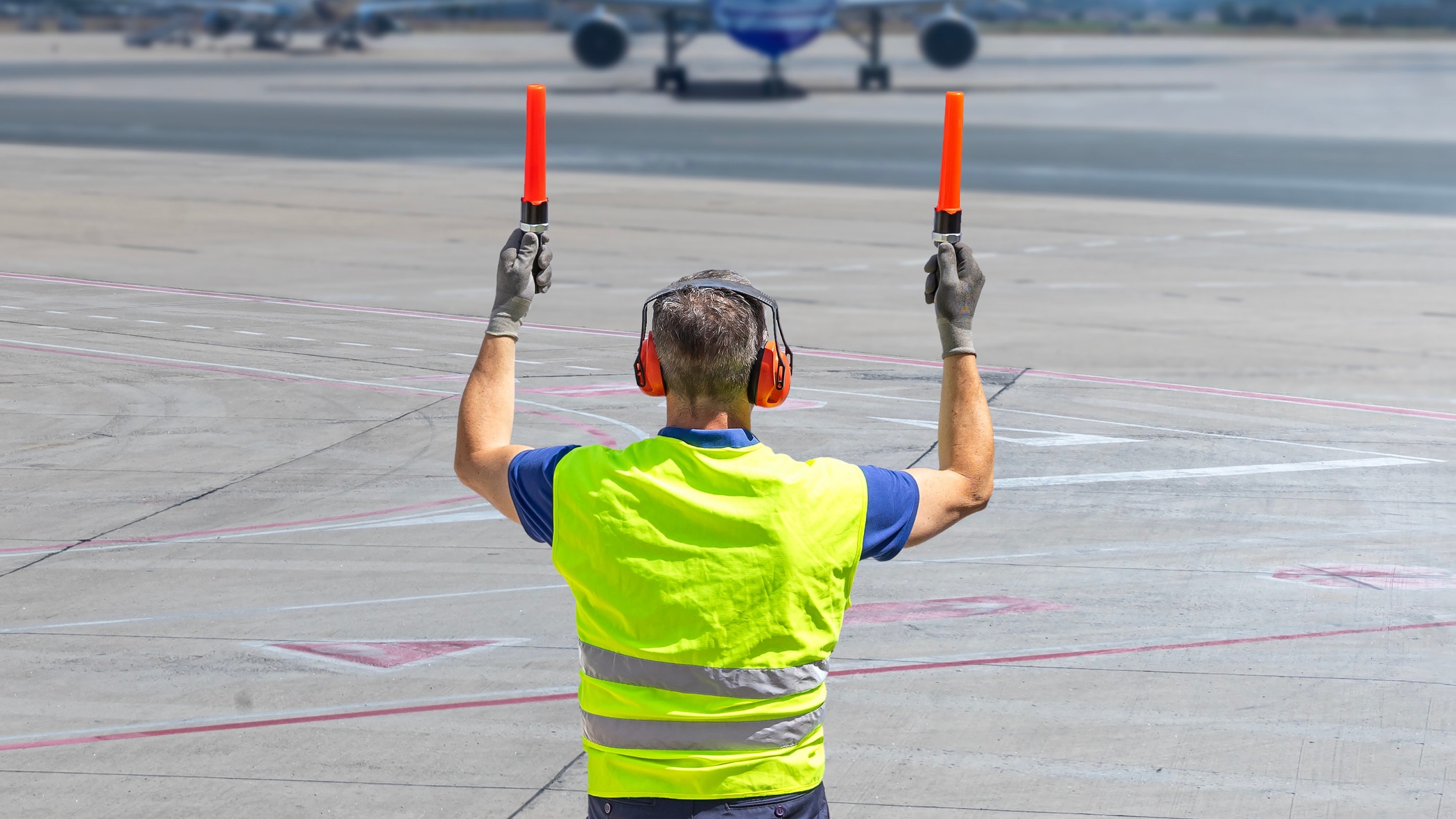 Rear view of airport marshaller in PPE guiding aircraft on runway with marshaling wands.