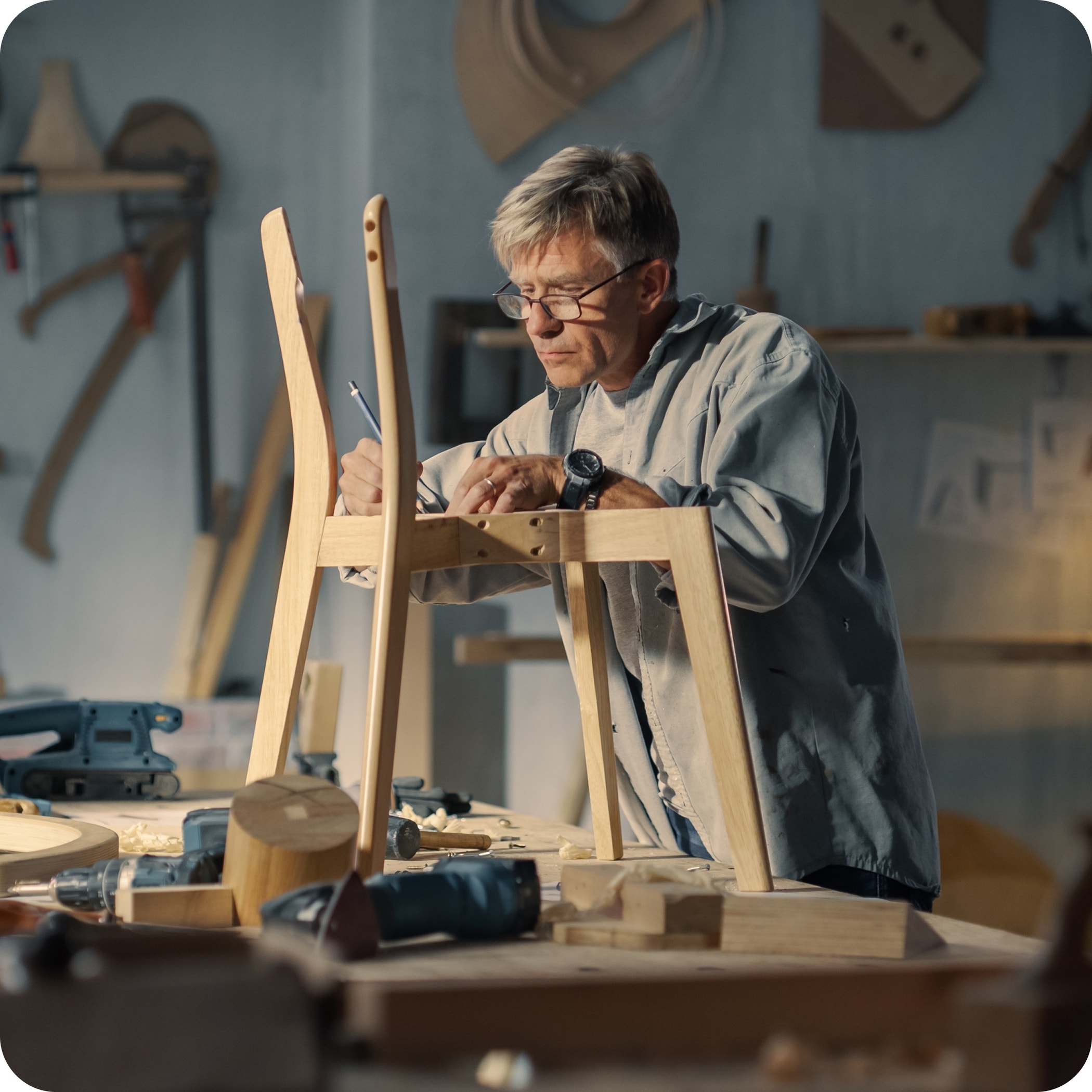 Middle aged carpenter putting on glasses, reading blueprints and starting to assemble parts of a wooden chair.
