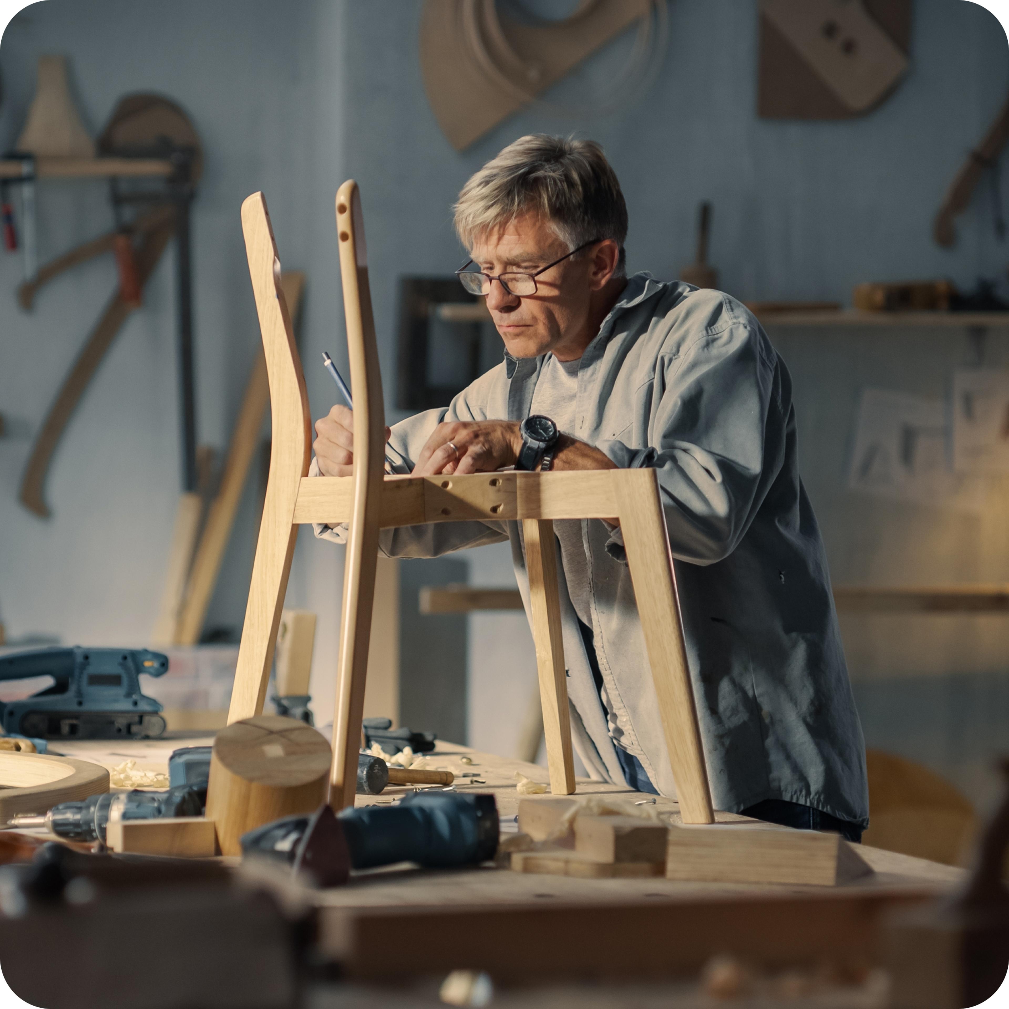 Middle aged carpenter putting on glasses, reading blueprints and starting to assemble parts of a wooden chair.