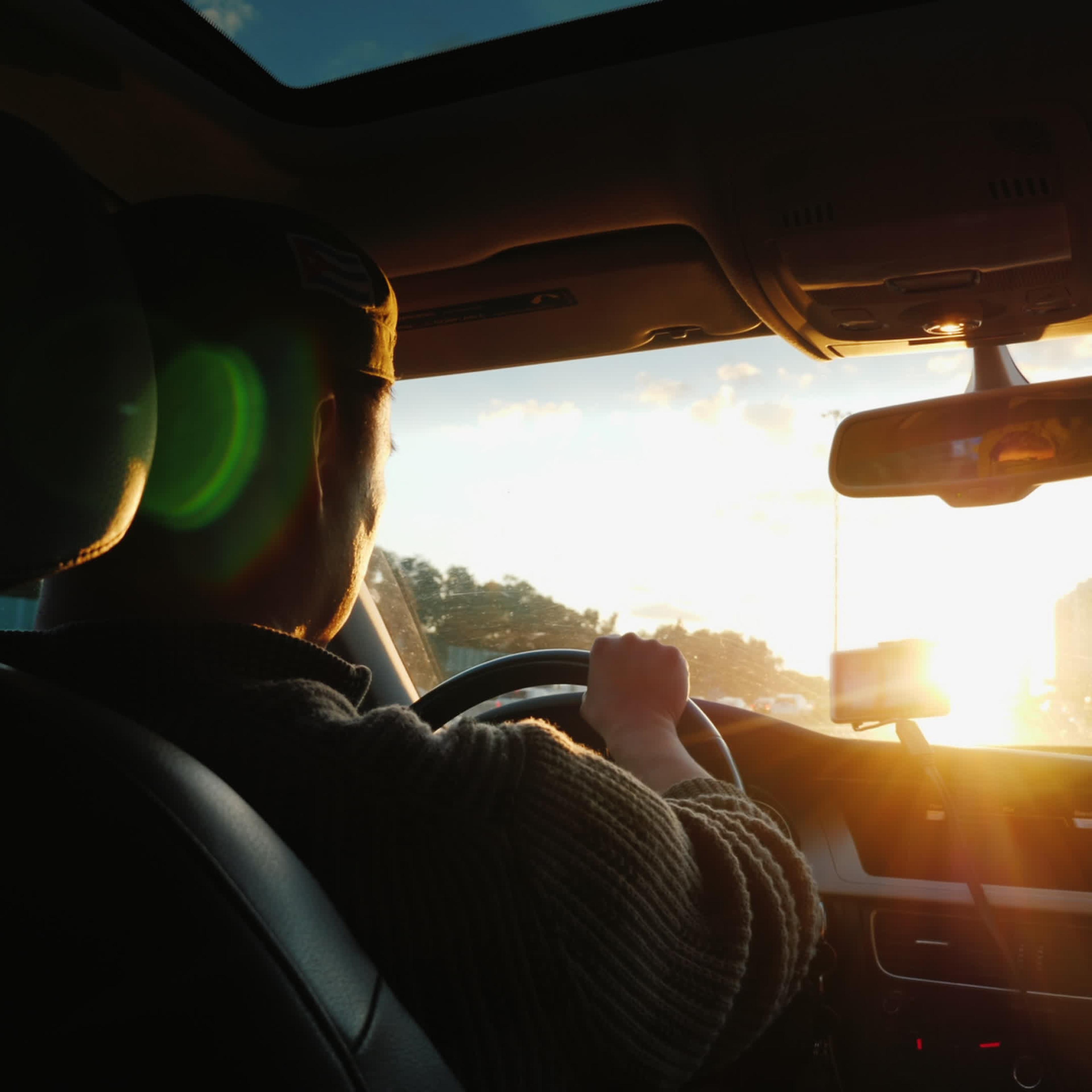 An Asian man drives a car along the highway, the setting sun shines in the windshield.