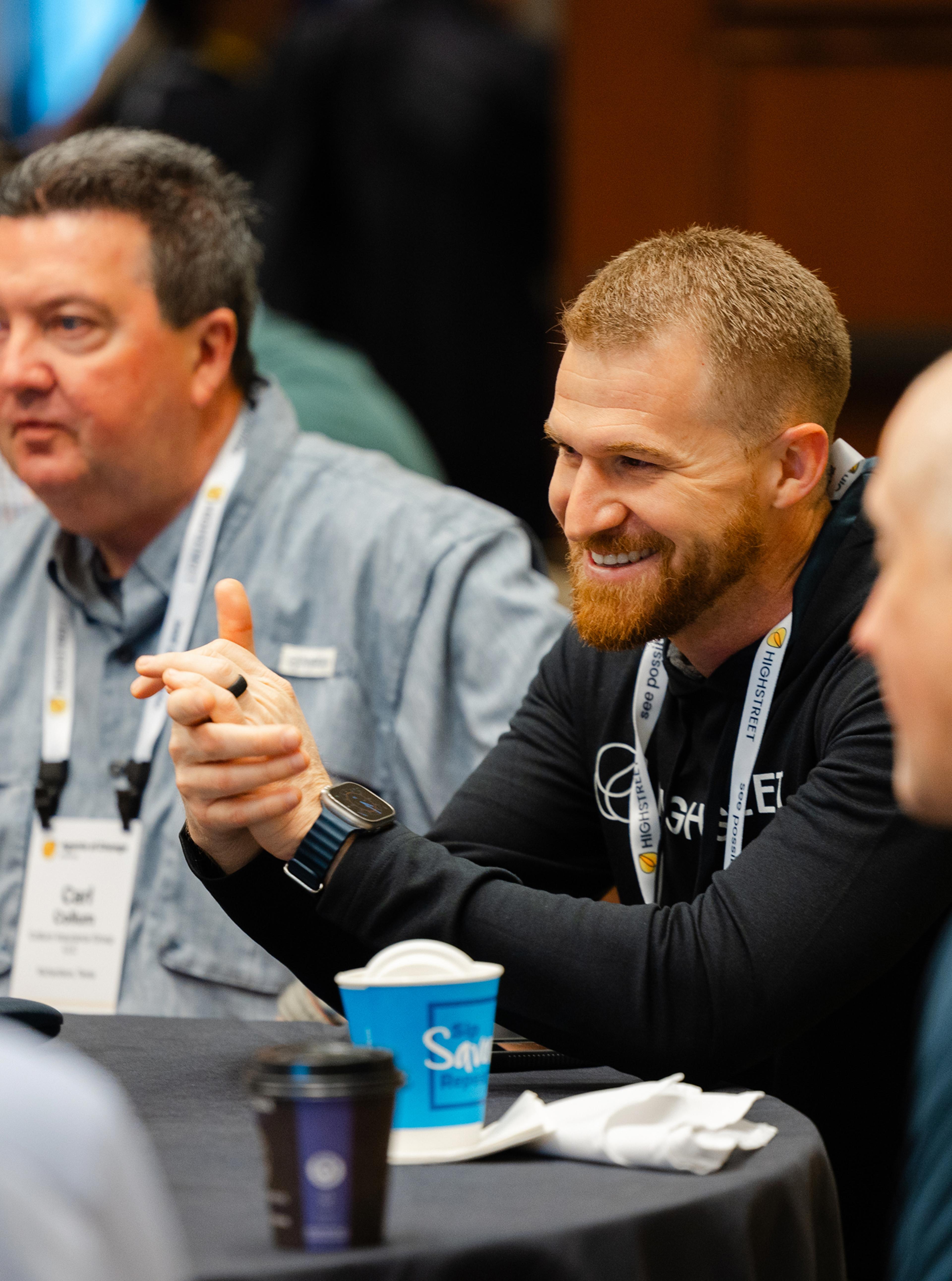 Highstreet red haired man with beard smiling and conversing at a table with other men.