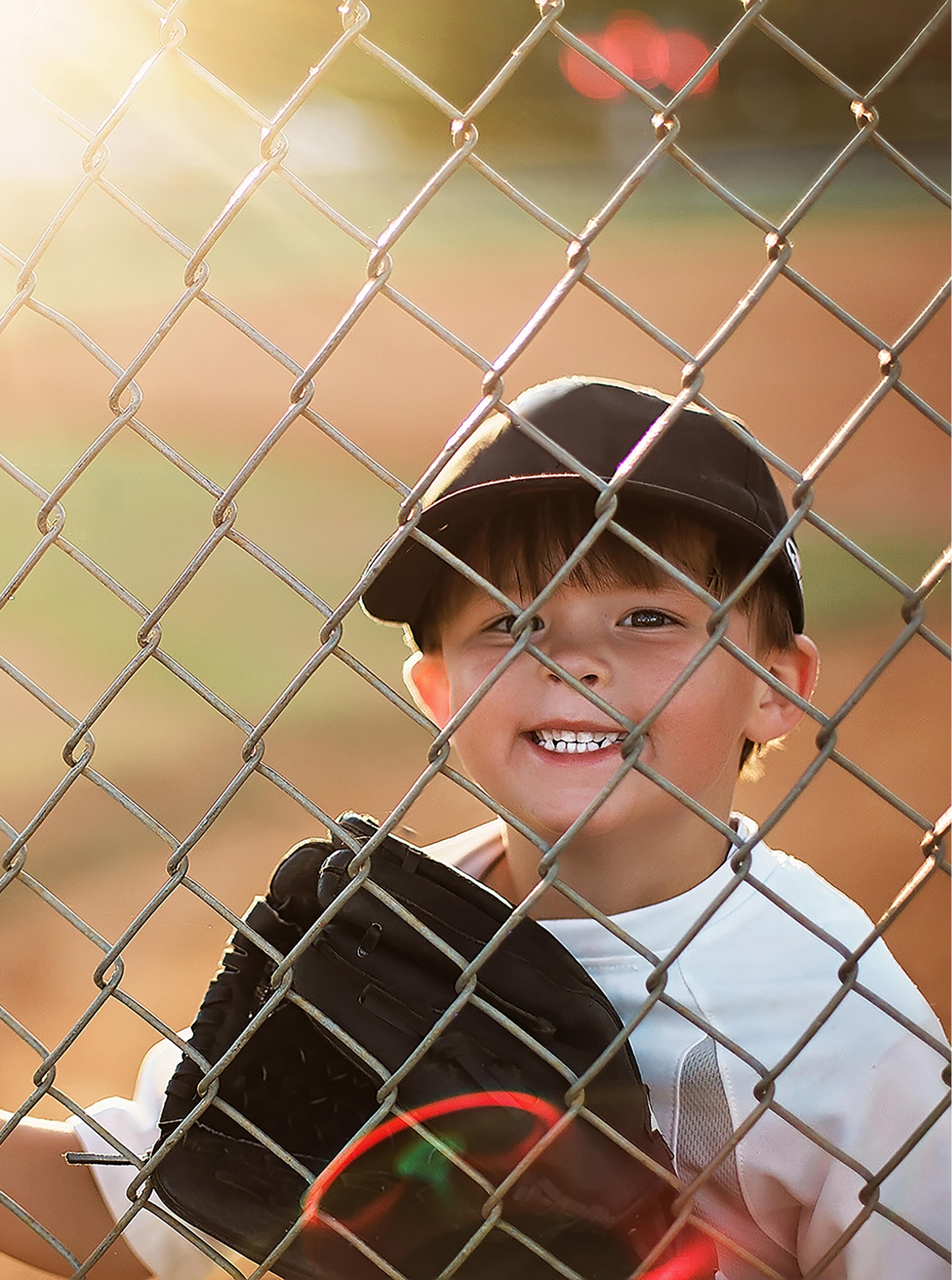 Little boy at a baseball game looking through the fence smiling.