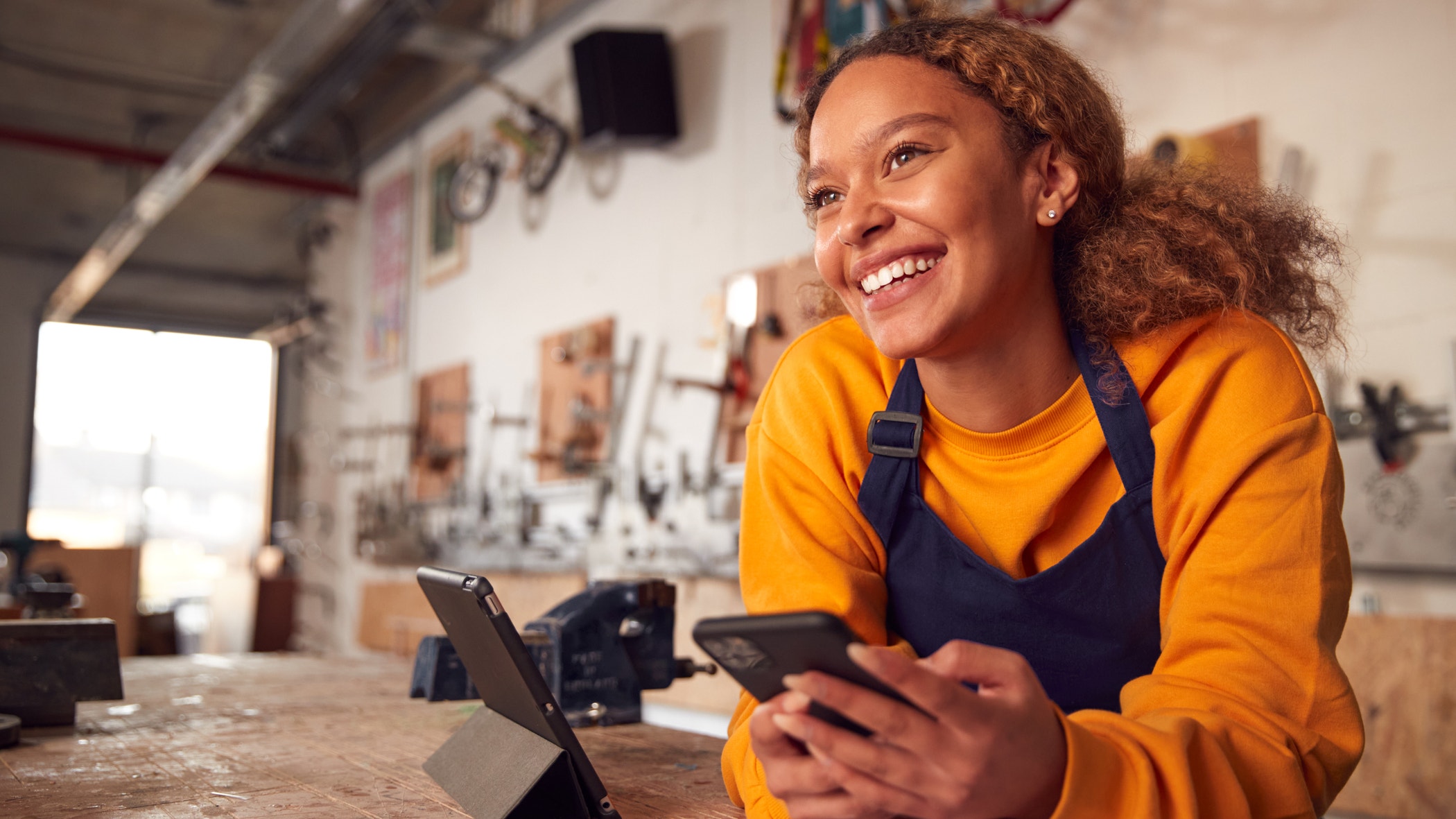 Female business owner In workshop using digital tablet and holding mobile phone.