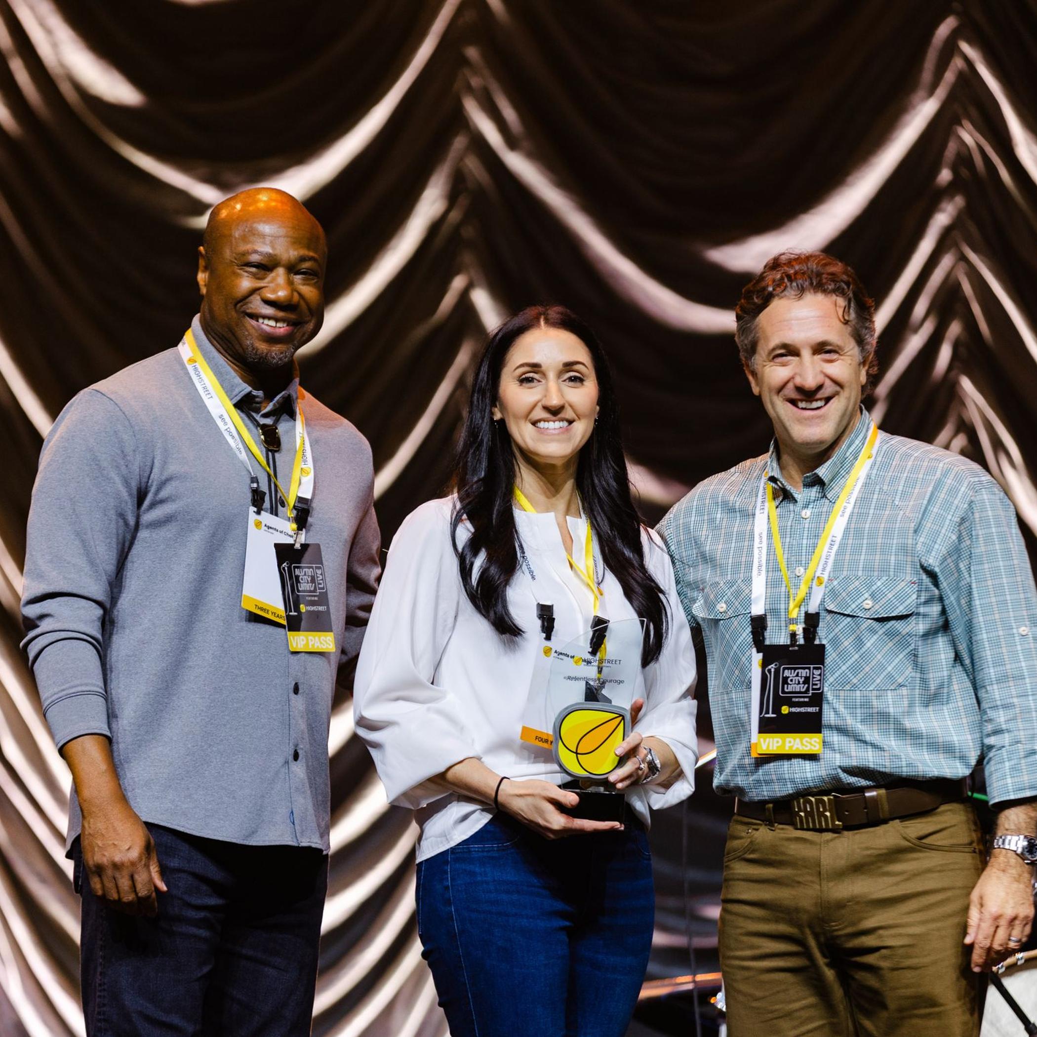 Three people smiling on stage during an awards ceremony, holding a yellow-accented trophy.