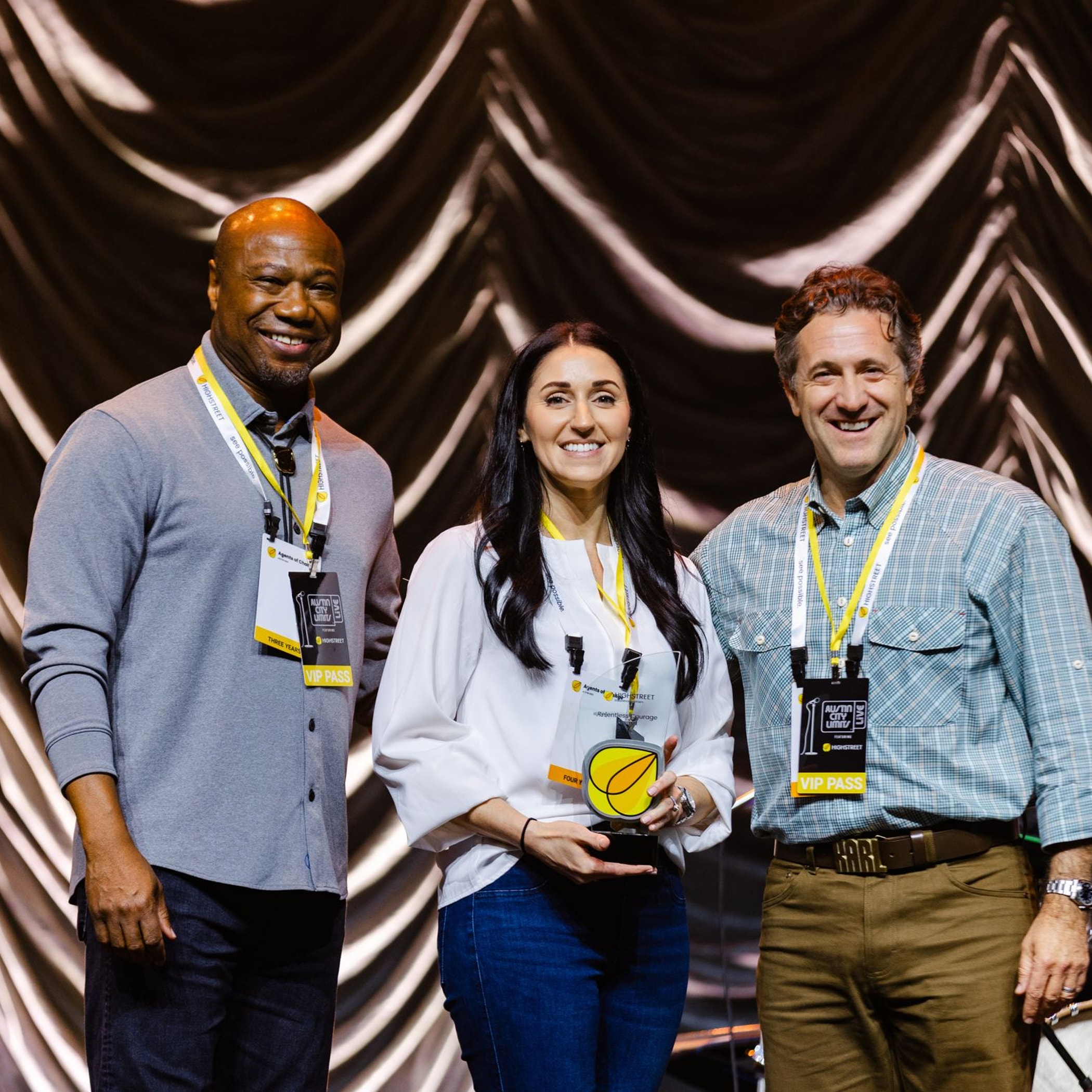 Three people smiling on stage during an awards ceremony, holding a yellow-accented trophy.