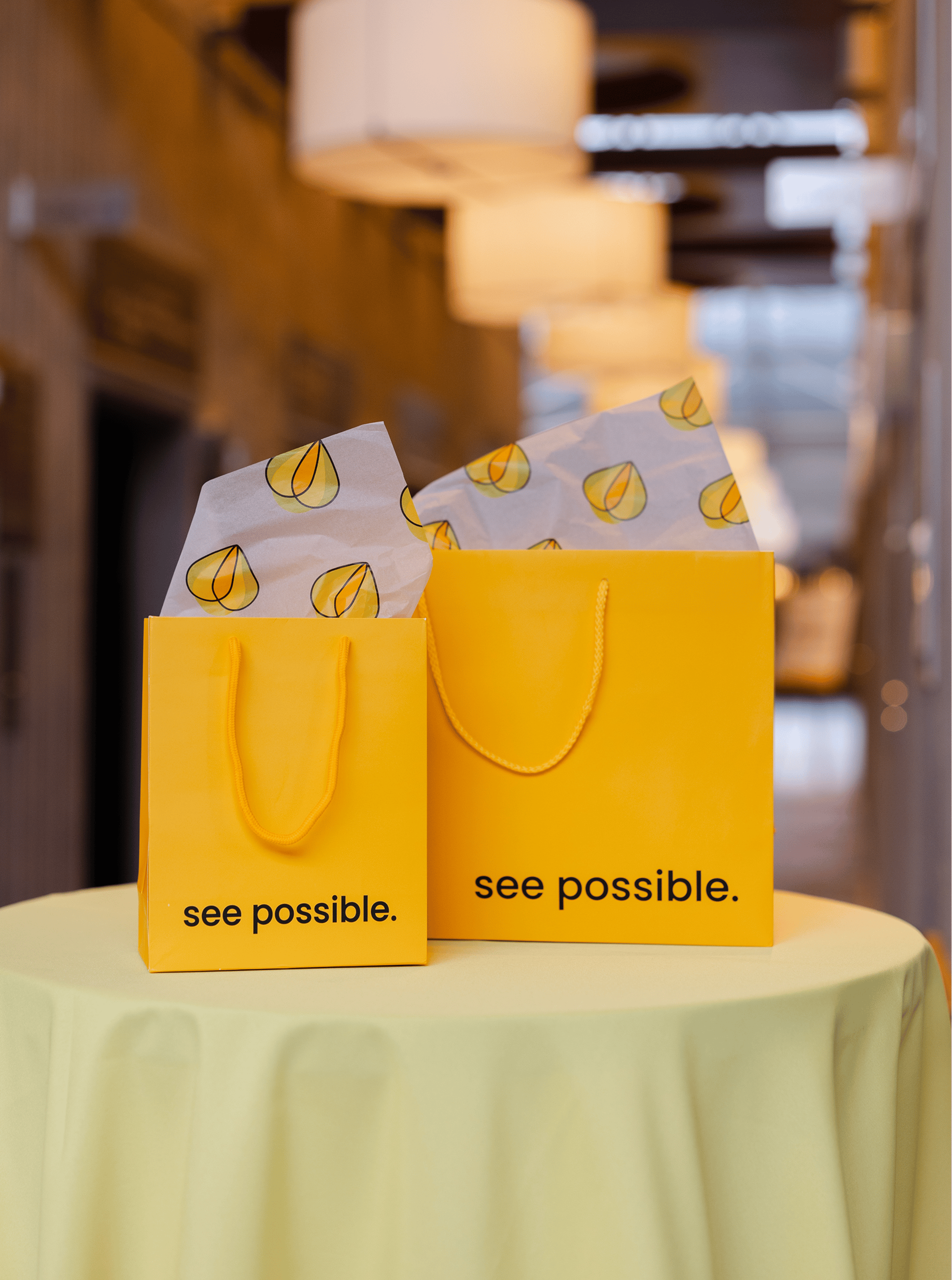 Two bright yellow gift bags with “see possible.” printed on them, placed on a round table with soft lighting in the background.
