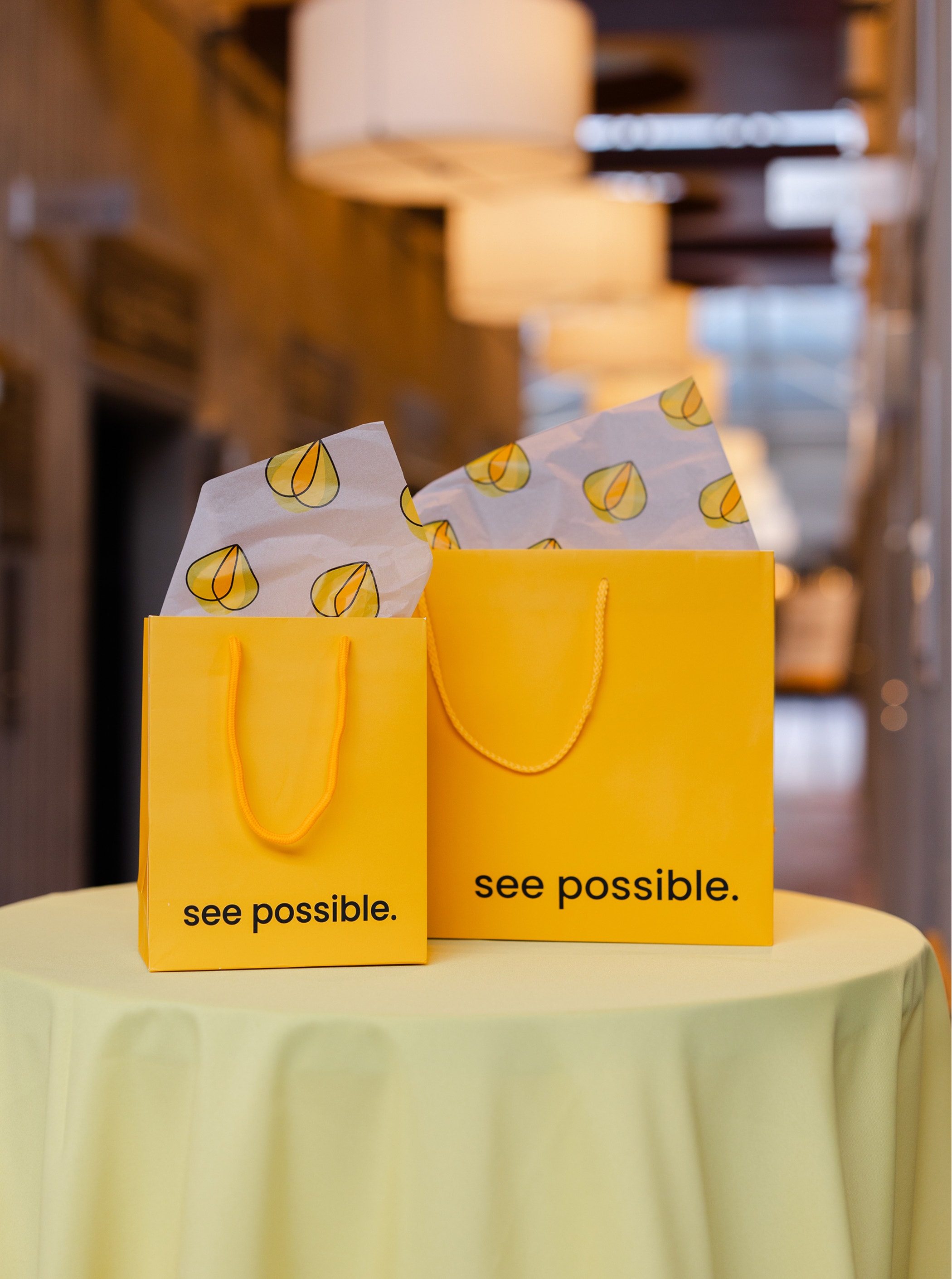 Two bright yellow gift bags with “see possible.” printed on them, placed on a round table with soft lighting in the background.