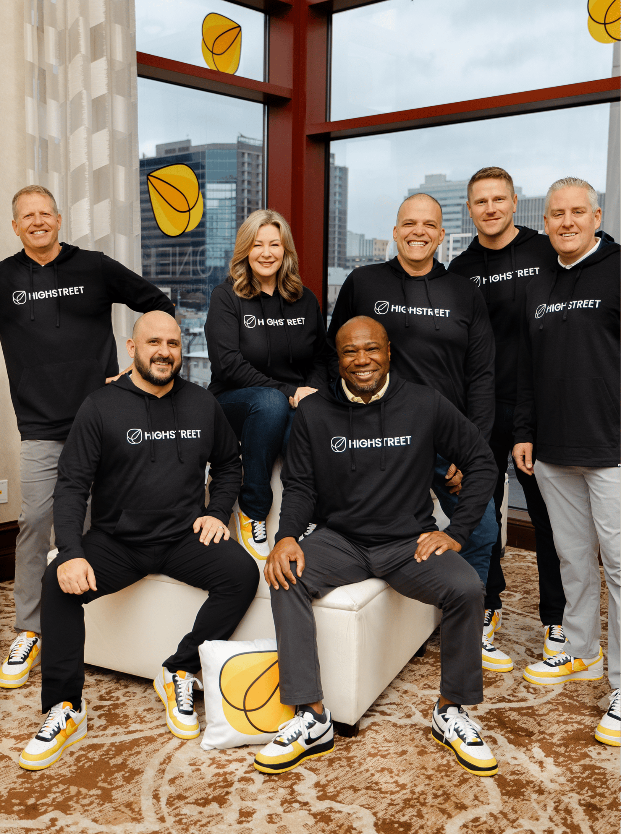 Group of people wearing matching Highstreet sweatshirts and yellow-accented sneakers, posing indoors with city buildings visible through large windows.
