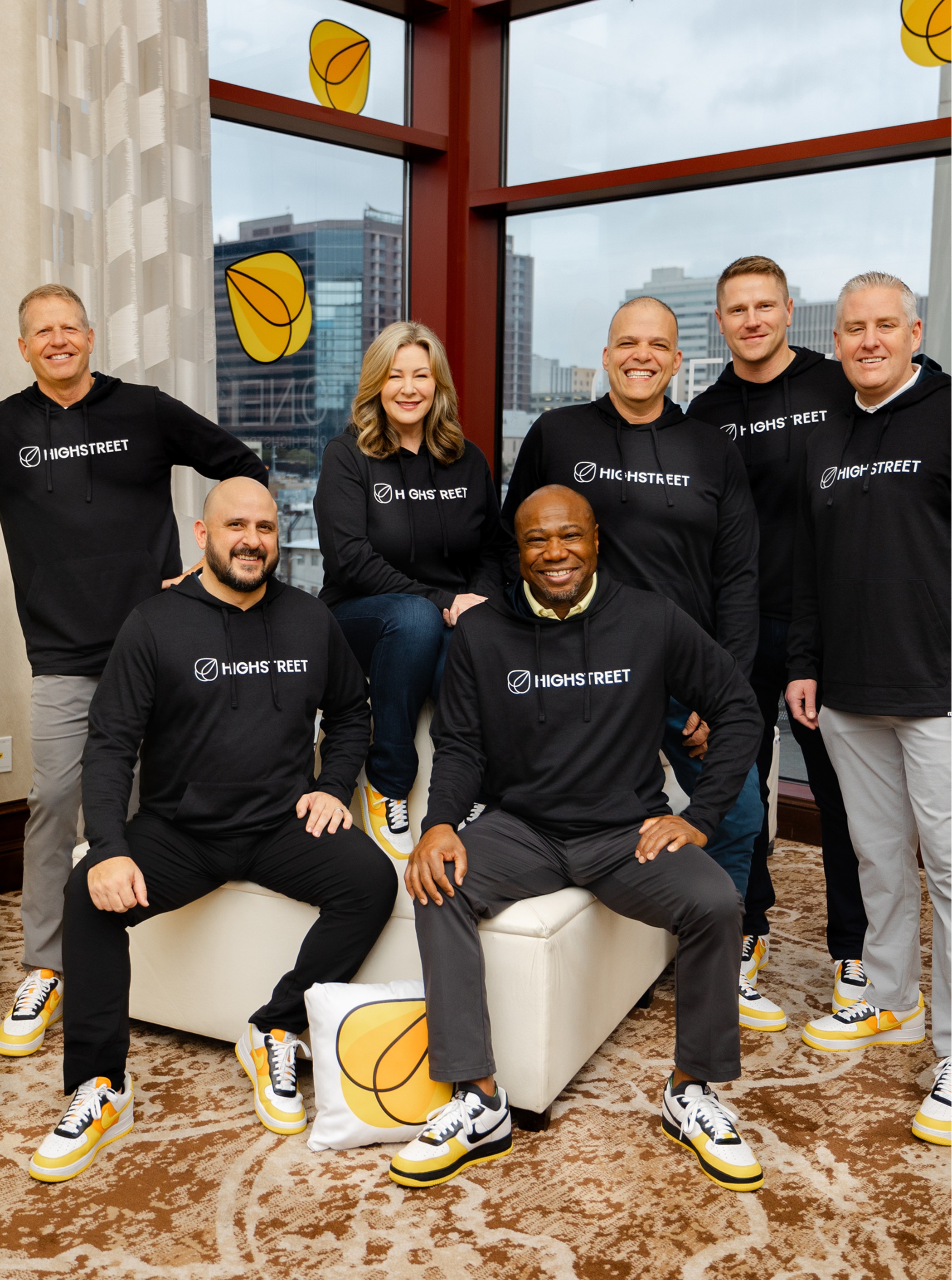 Group of people wearing matching Highstreet sweatshirts and yellow-accented sneakers, posing indoors with city buildings visible through large windows.