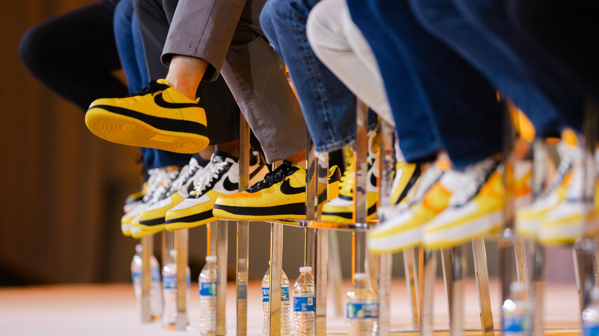 Close-up of people sitting in a row wearing matching yellow and black sneakers, with water bottles lined up on the floor beneath their chairs.