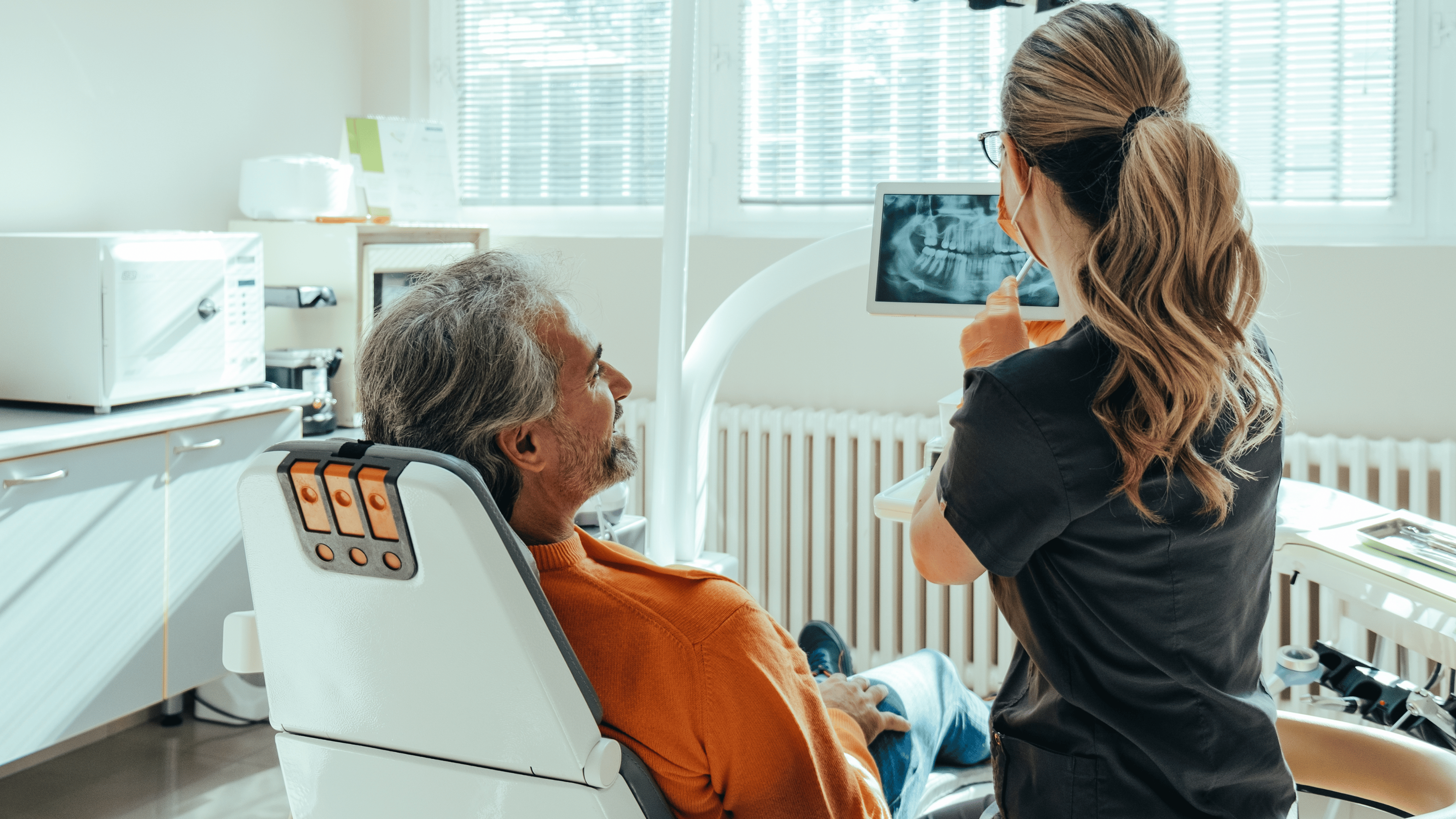 An anonymous female dentist explaining teeth x-ray on a digital tablet to smiling mature patient at dentist's office.