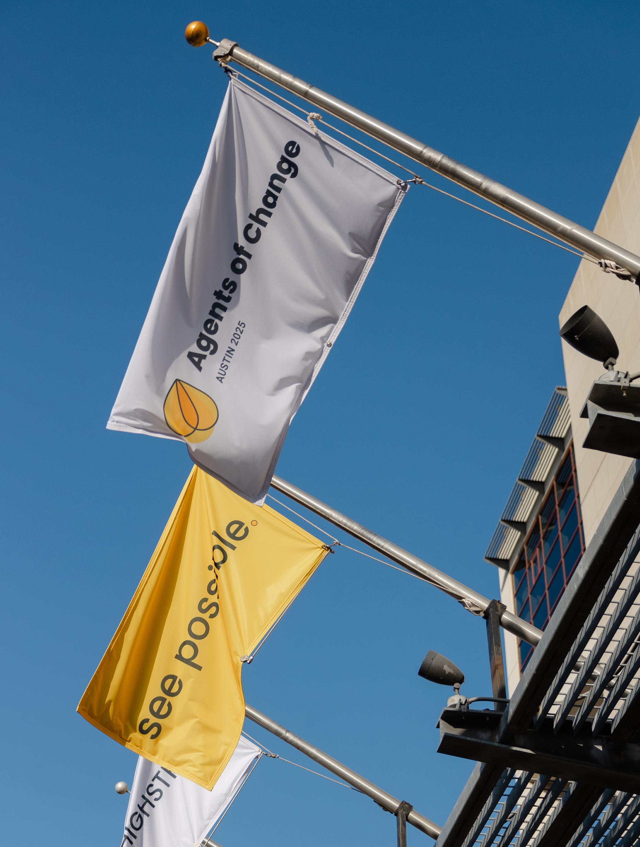 Flags waving outdoors under a clear blue sky, one reading “Agents of Change Austin 2025” and another yellow one reading “see possible.”