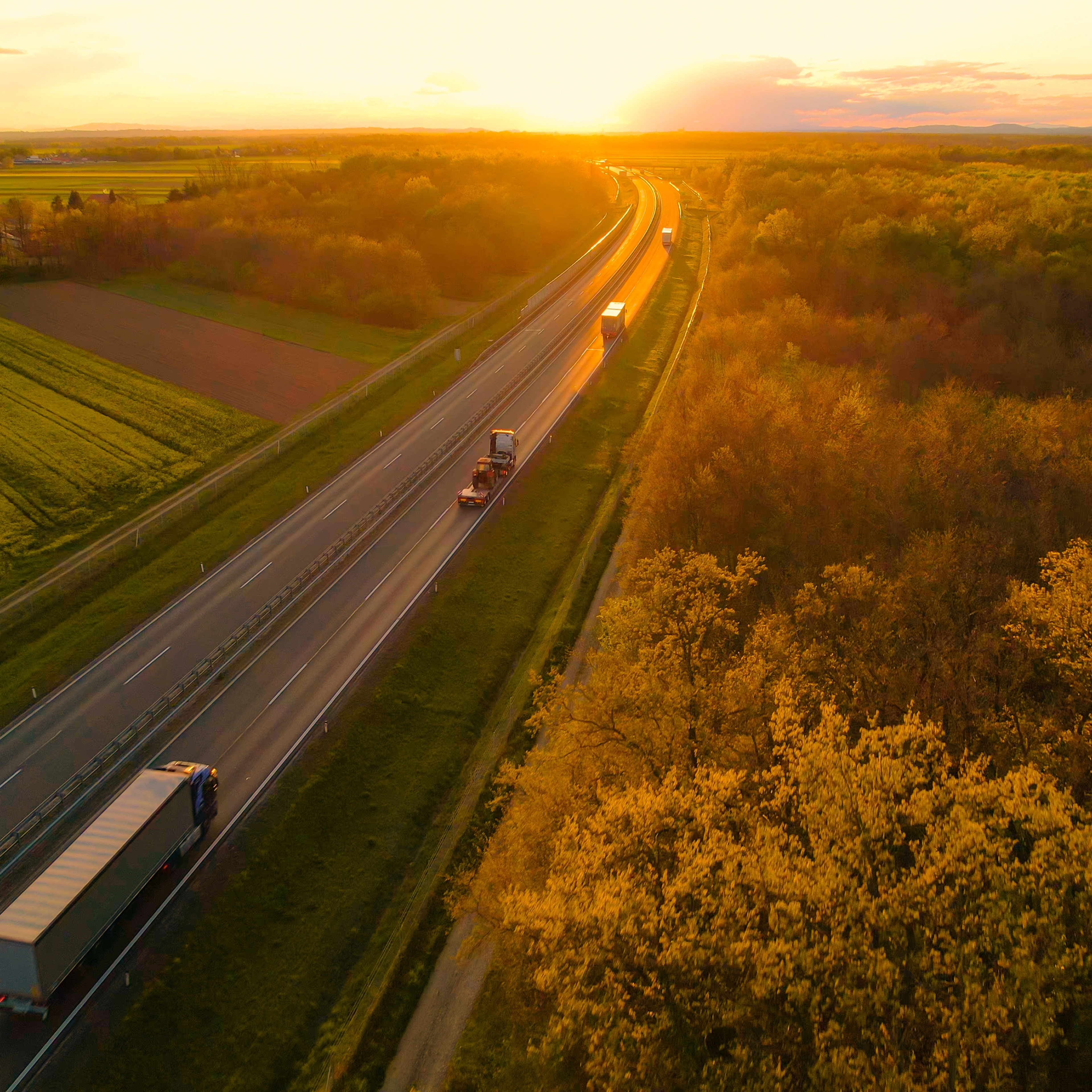 Aerial view of cargo trucks driving on a countryside highway in golden light, transporting goods through a scenic landscape.