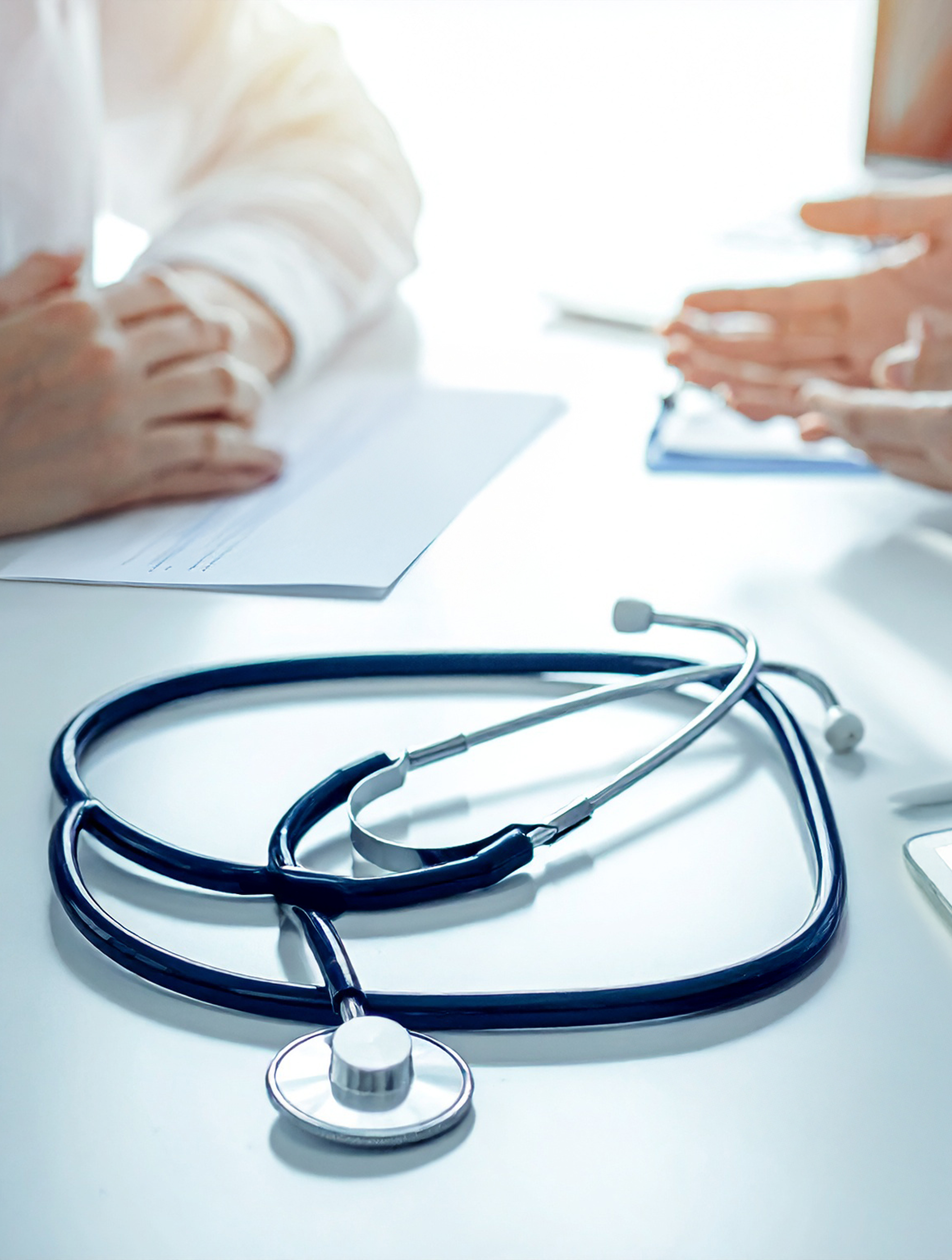 Stethoscope on a desk with two people talking in the background during a medical consultation.