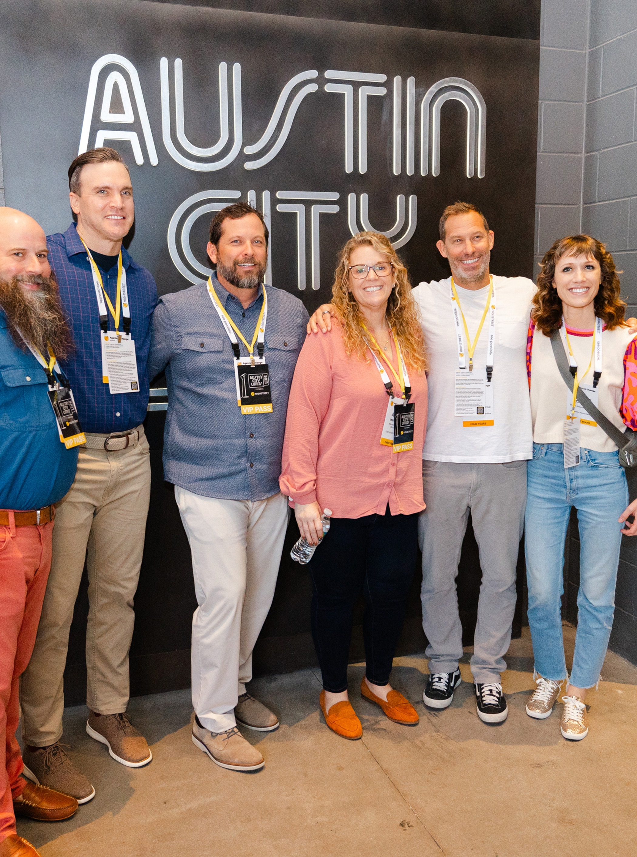 Group of people standing together and smiling in front of a black wall with large white letters reading “Austin City.”
