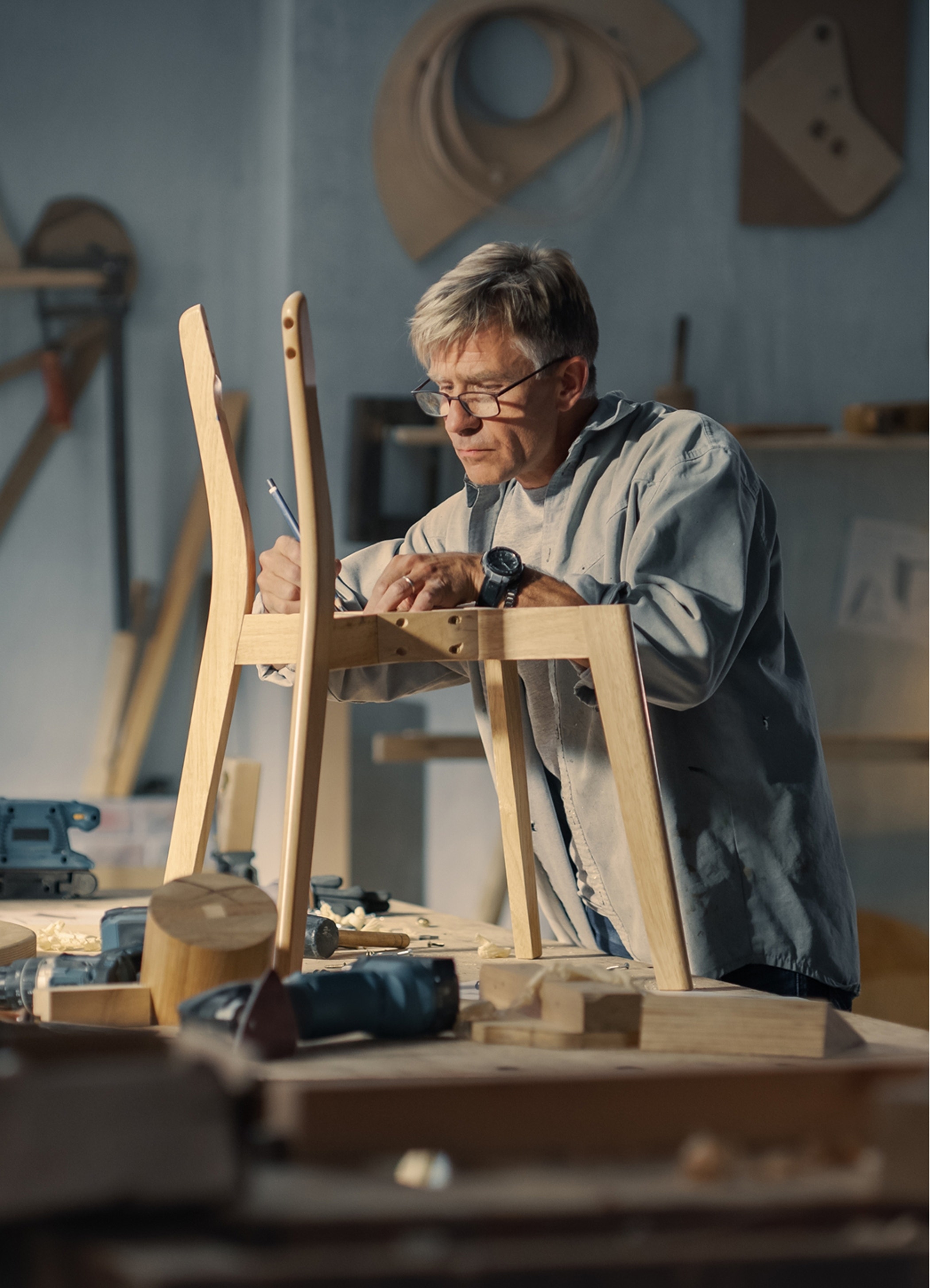 Middle aged carpenter putting on glasses, reading blueprints and starting to assemble parts of a wooden chair.