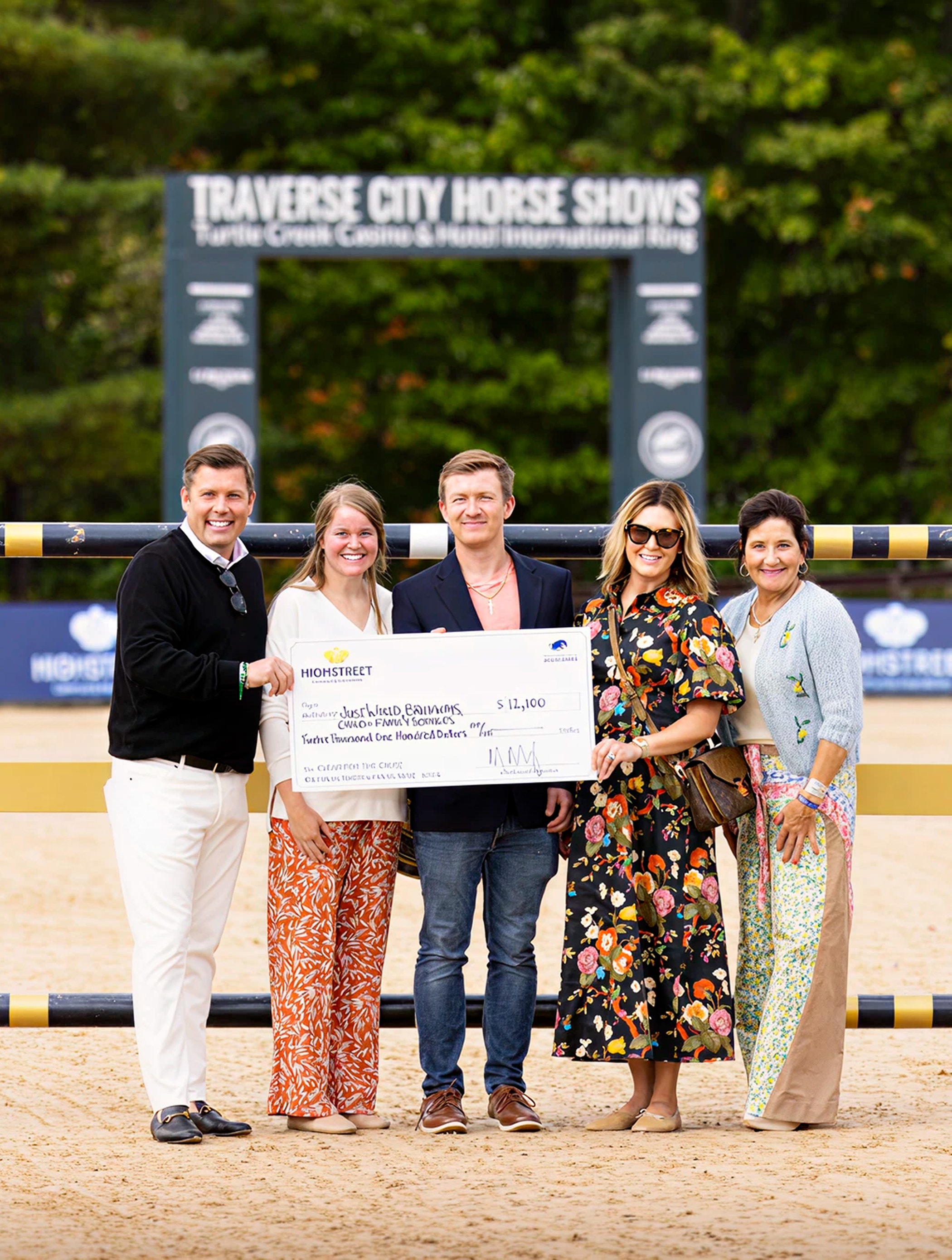 Group of people standing outdoors holding a large donation check during a horse show event, with the “Traverse City Horse Shows” sign visible in the background.