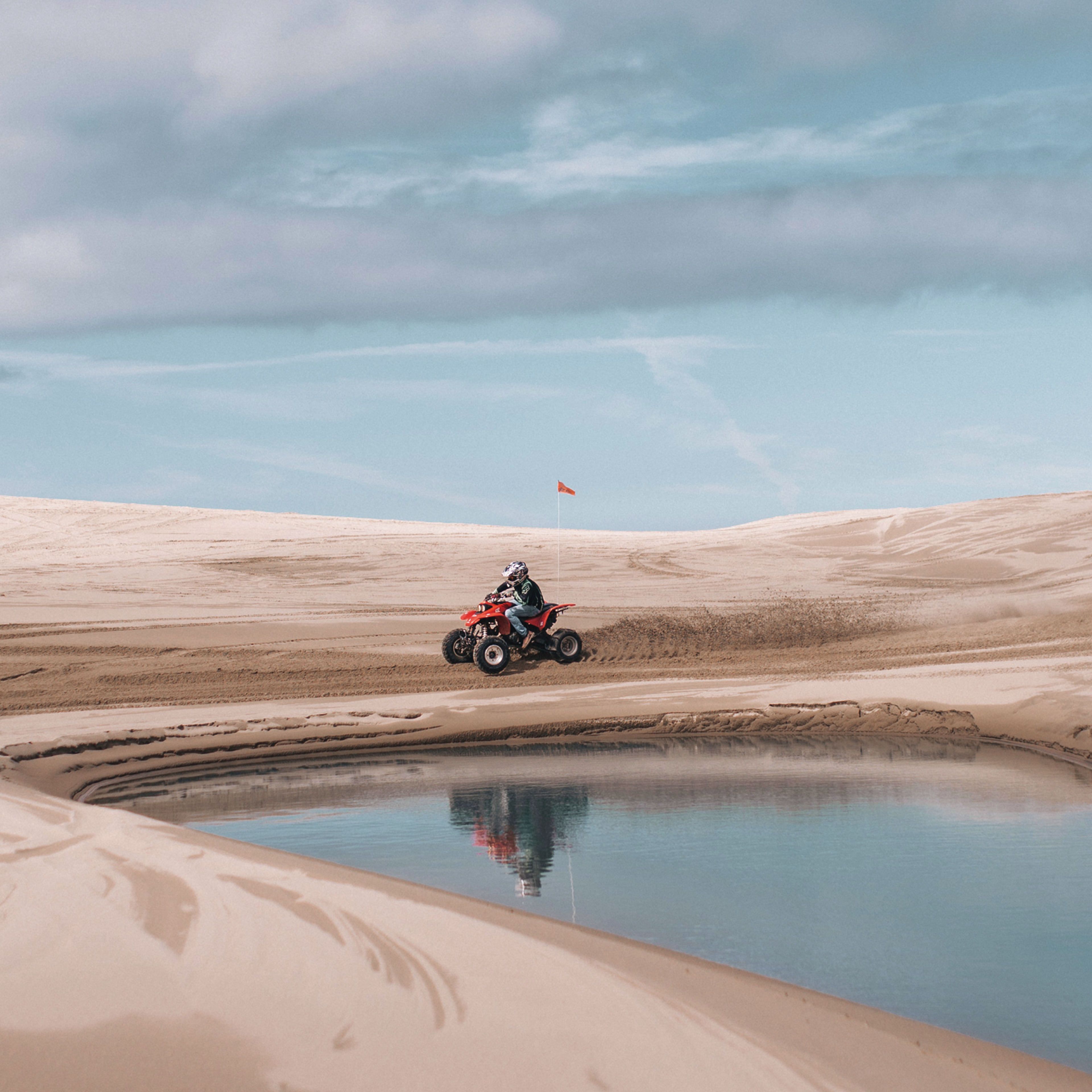 Person riding a red ATV on sandy dunes near a reflective water pool under a blue sky.