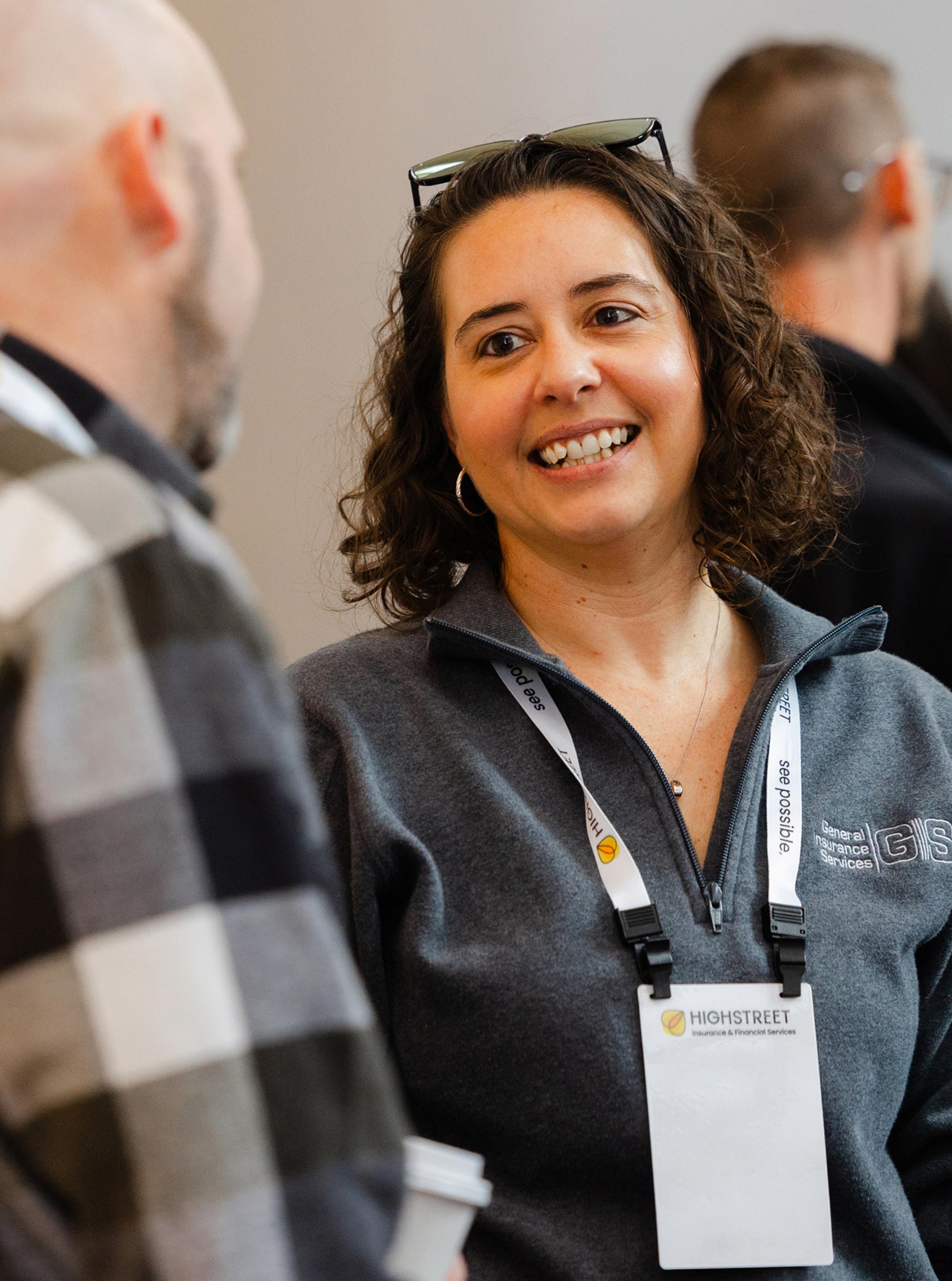 Woman wearing a conference badge and a gray Highstreet jacket smiling while talking to a colleague at an event.
