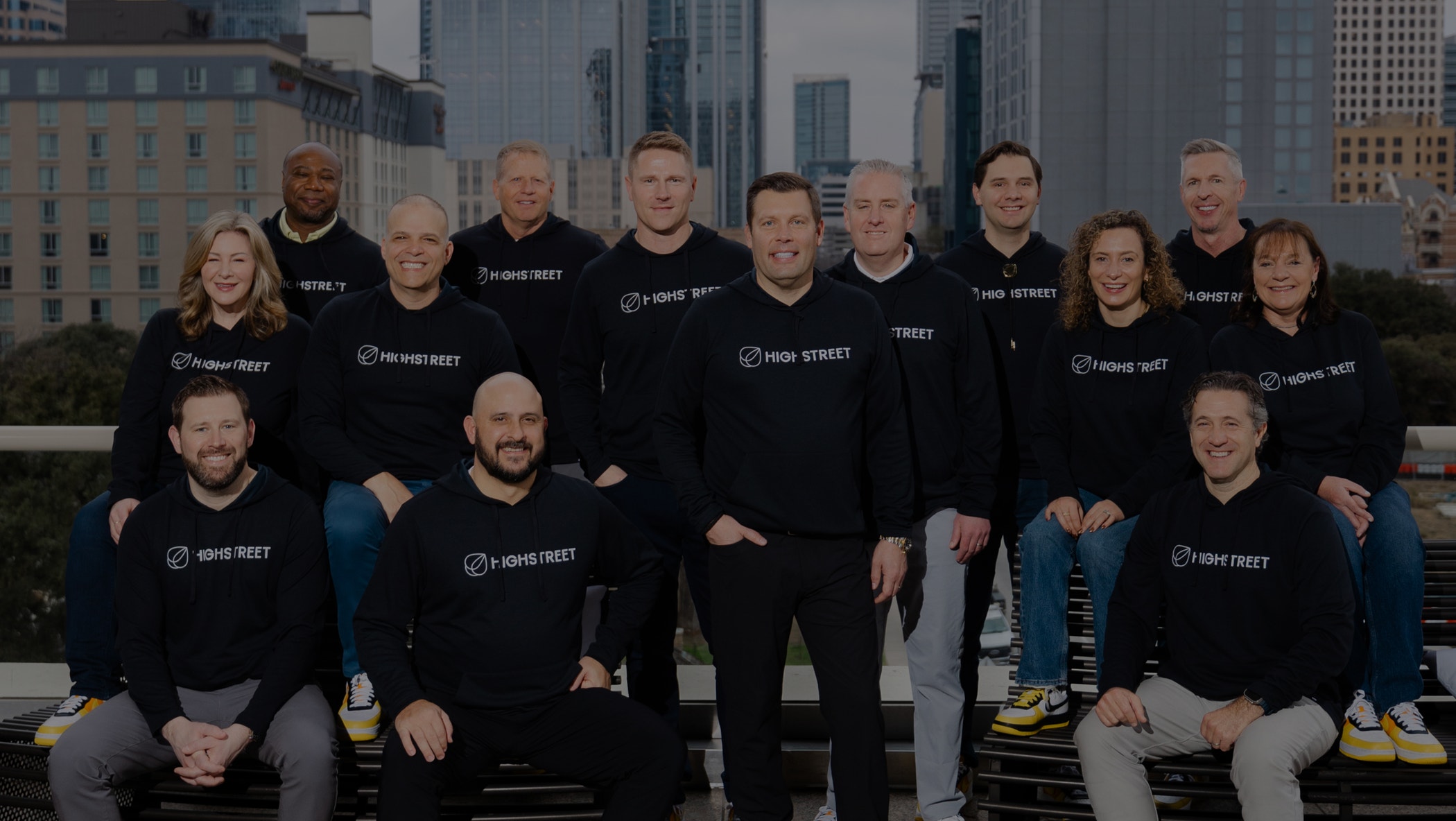 Group of people wearing matching Highstreet sweatshirts and yellow-accented sneakers, posing indoors with city buildings visible through large windows.