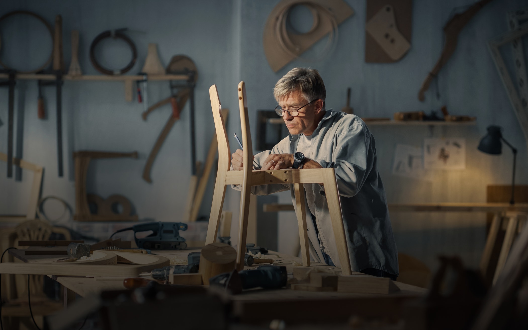 Middle aged carpenter putting on glasses, reading blueprints and starting to assemble parts of a wooden chair.