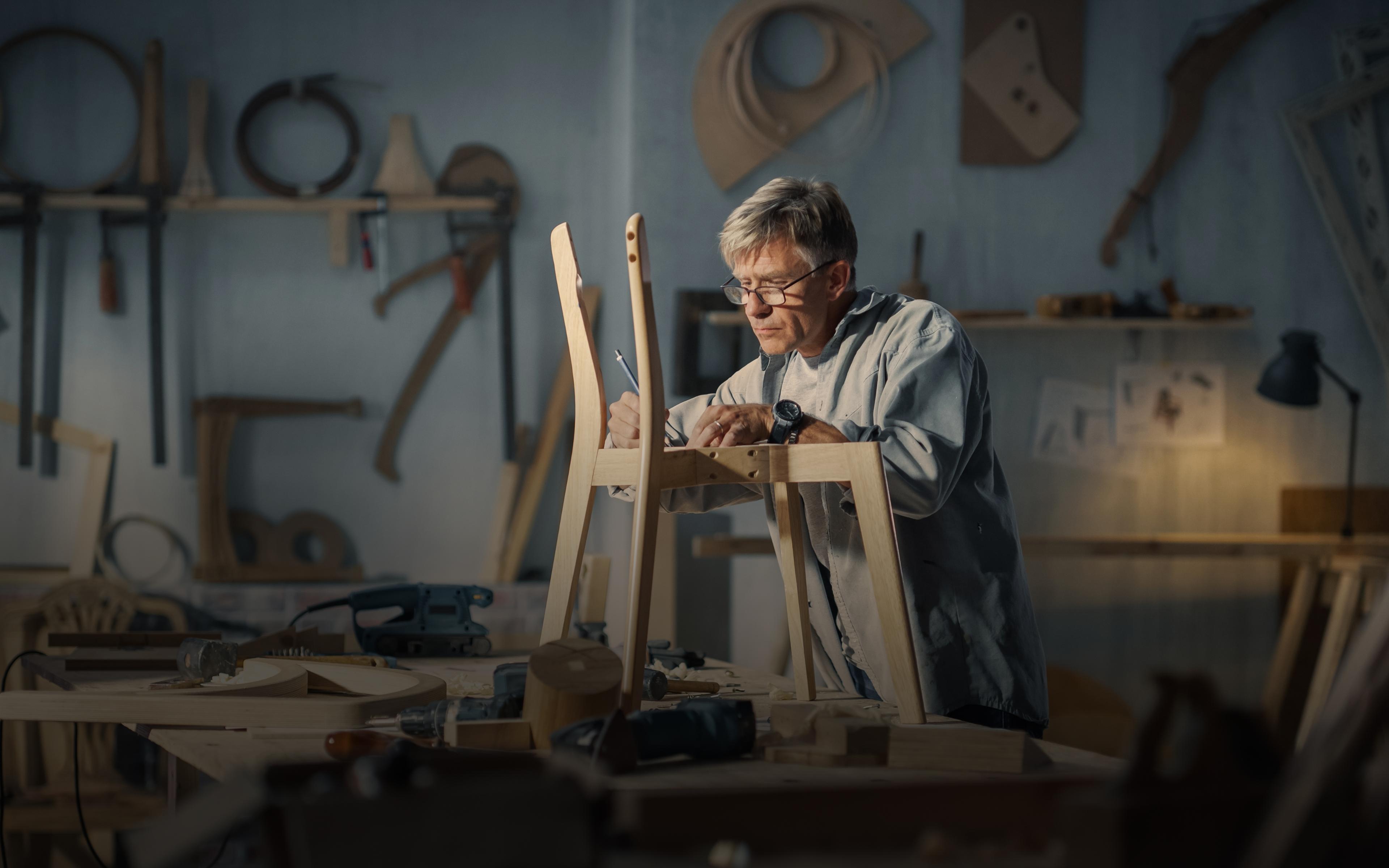 Middle aged carpenter putting on glasses, reading blueprints and starting to assemble parts of a wooden chair.