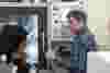A man in a plaid shirt talks to a woman with dark hair in front of a Haas CNC machine in a factory setting. The man holds the machine’s control handle while the woman listens attentively.
