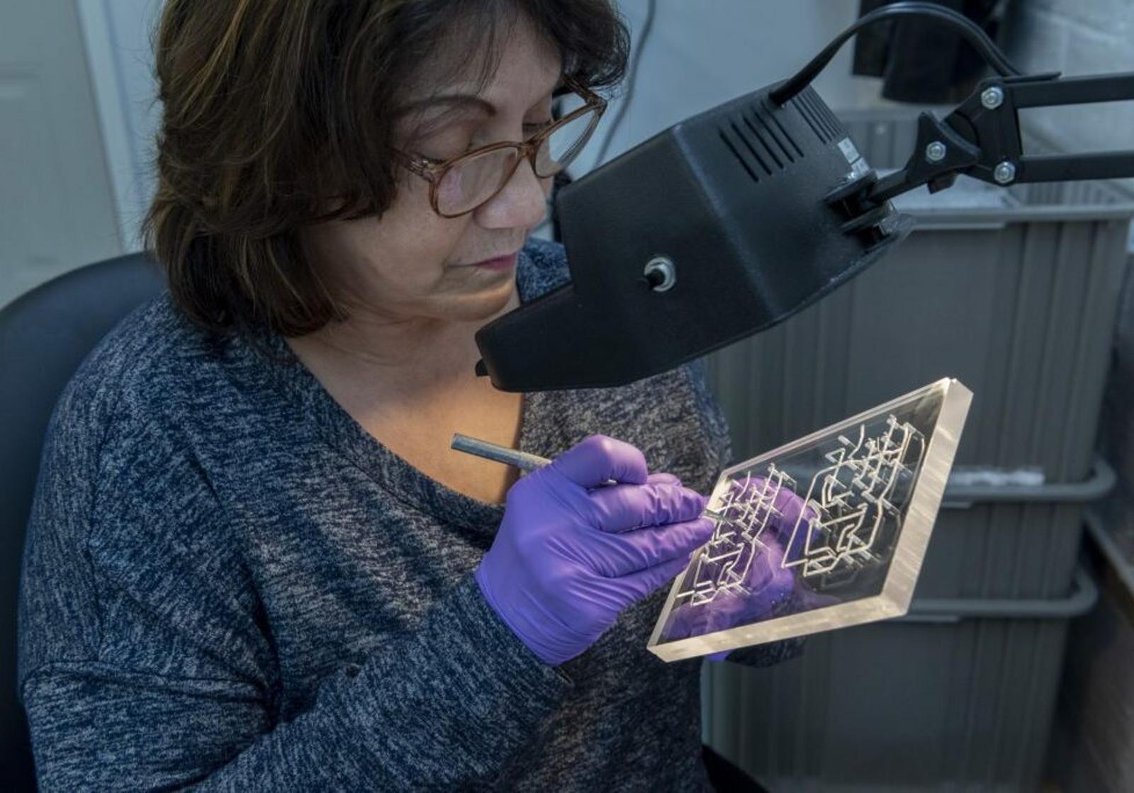 A woman wearing glasses and purple gloves uses a tool to work on a transparent plastic manifold with intricate designs, seated under a magnifying lamp in a workspace.