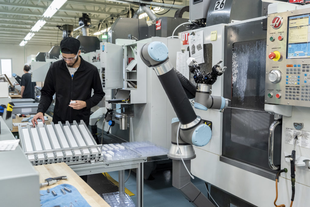 A man in a black jacket and cap works beside a robotic arm and CNC machines in a modern factory, handling small plastic parts on a tray under bright industrial lighting.