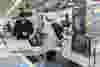 A man in a black jacket and cap works beside a robotic arm and CNC machines in a modern factory, handling small plastic parts on a tray under bright industrial lighting.