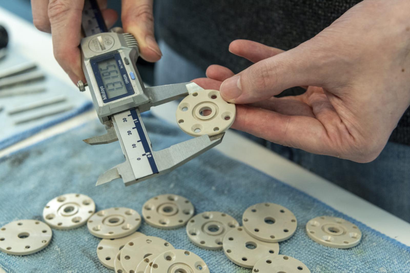 A person uses digital calipers to measure the diameter of a round, beige PEEK plastic part with holes, while several similar parts are spread out on a blue cloth below.