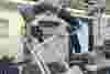 A robotic arm operates as part of a CNC machine, manipulating plastic manifolds held by blue clamps labeled Kurt. 