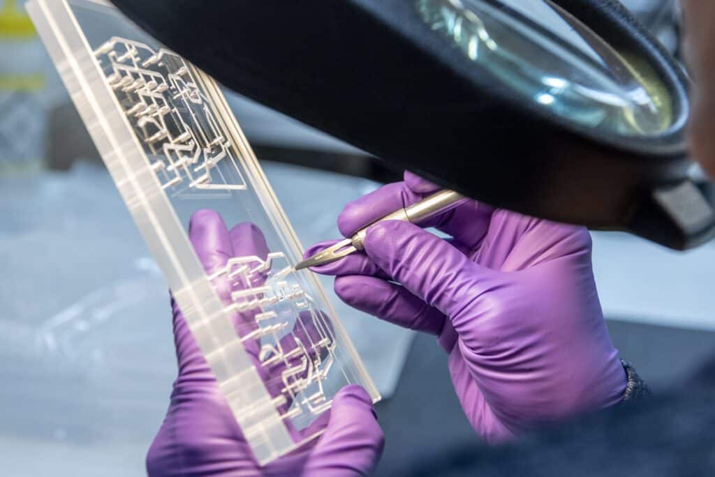 A person wearing purple gloves uses a metal tool to work on a transparent plastic fluidic manifold under a magnifying glass.