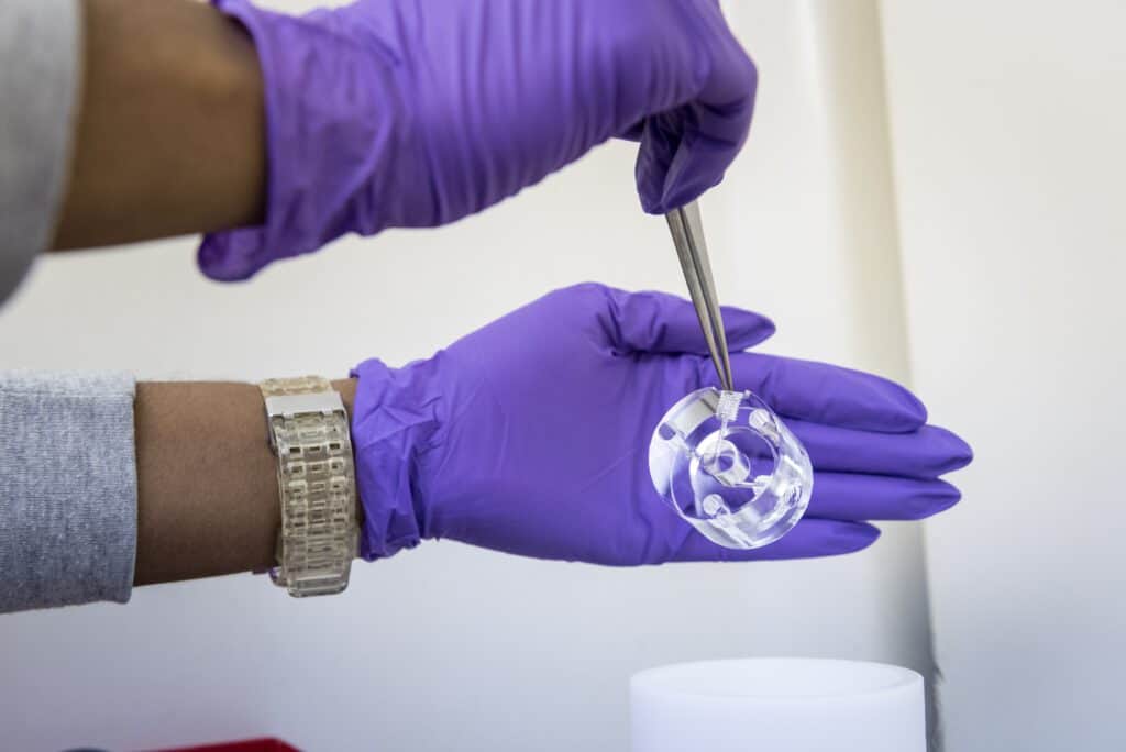 A person wearing a watch and purple gloves holds a small clear polished plastic component with tweezers in one hand and supports it with their other hand.