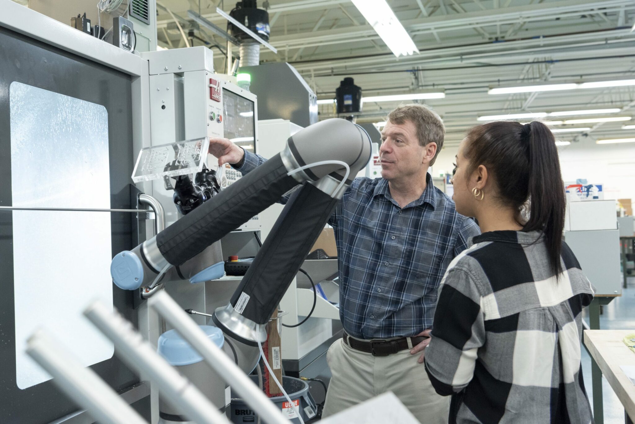 A man and a woman stand next to a CNC robotic arm in a modern factory holding a plastic manifold. The man points at a control panel while explaining something to the woman, who listens attentively.